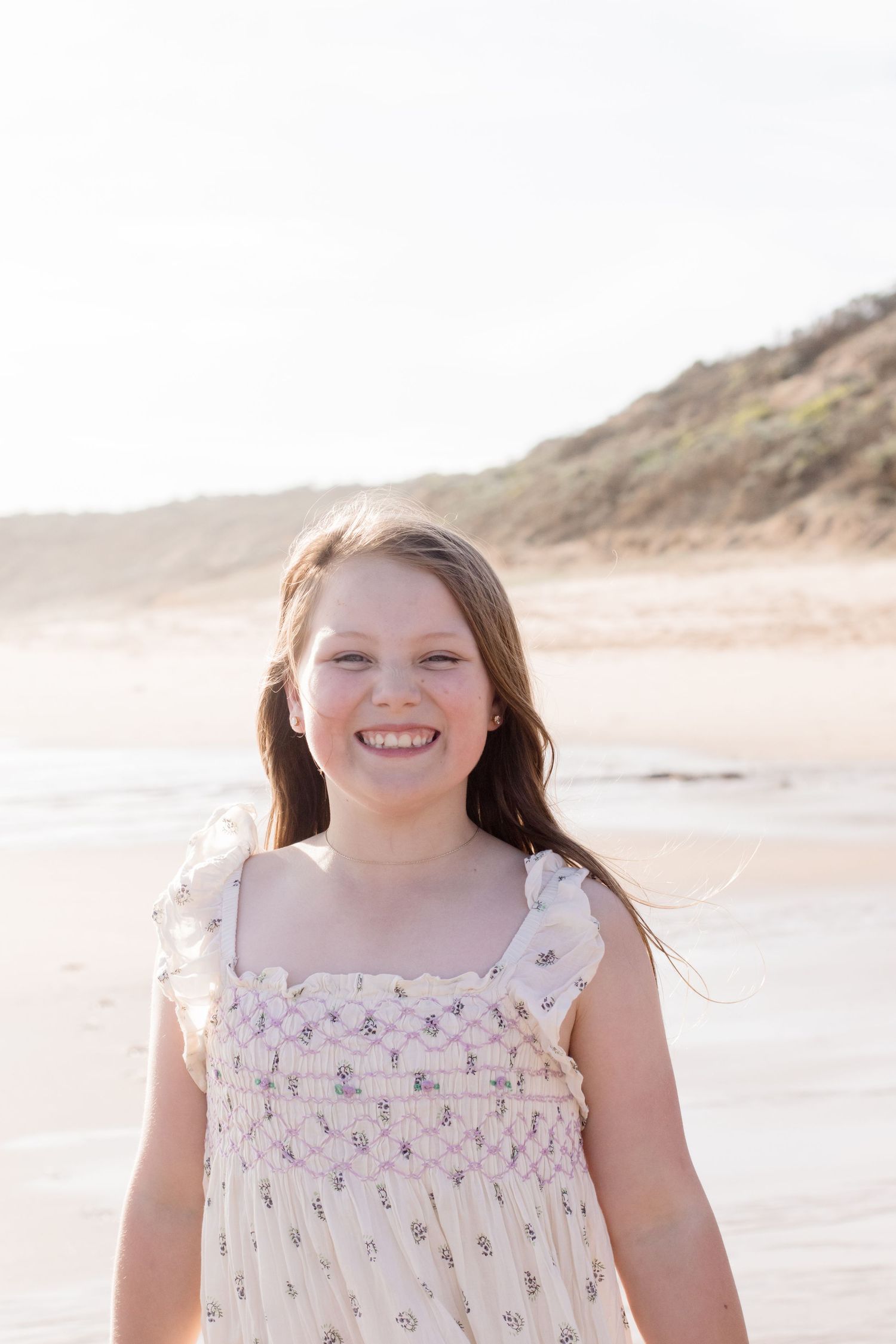 Portrait of someone in a floral dress smiling on a sunny beach with sand dunes in the background.