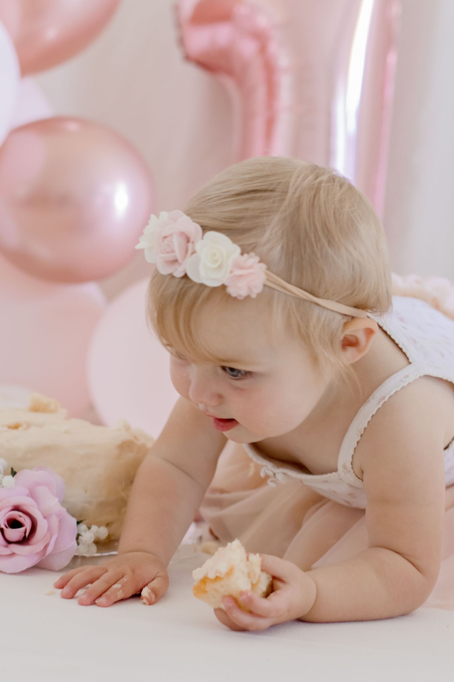 A small toddler in a white dress with floral headband explores pink decor and balloons at a party setup.