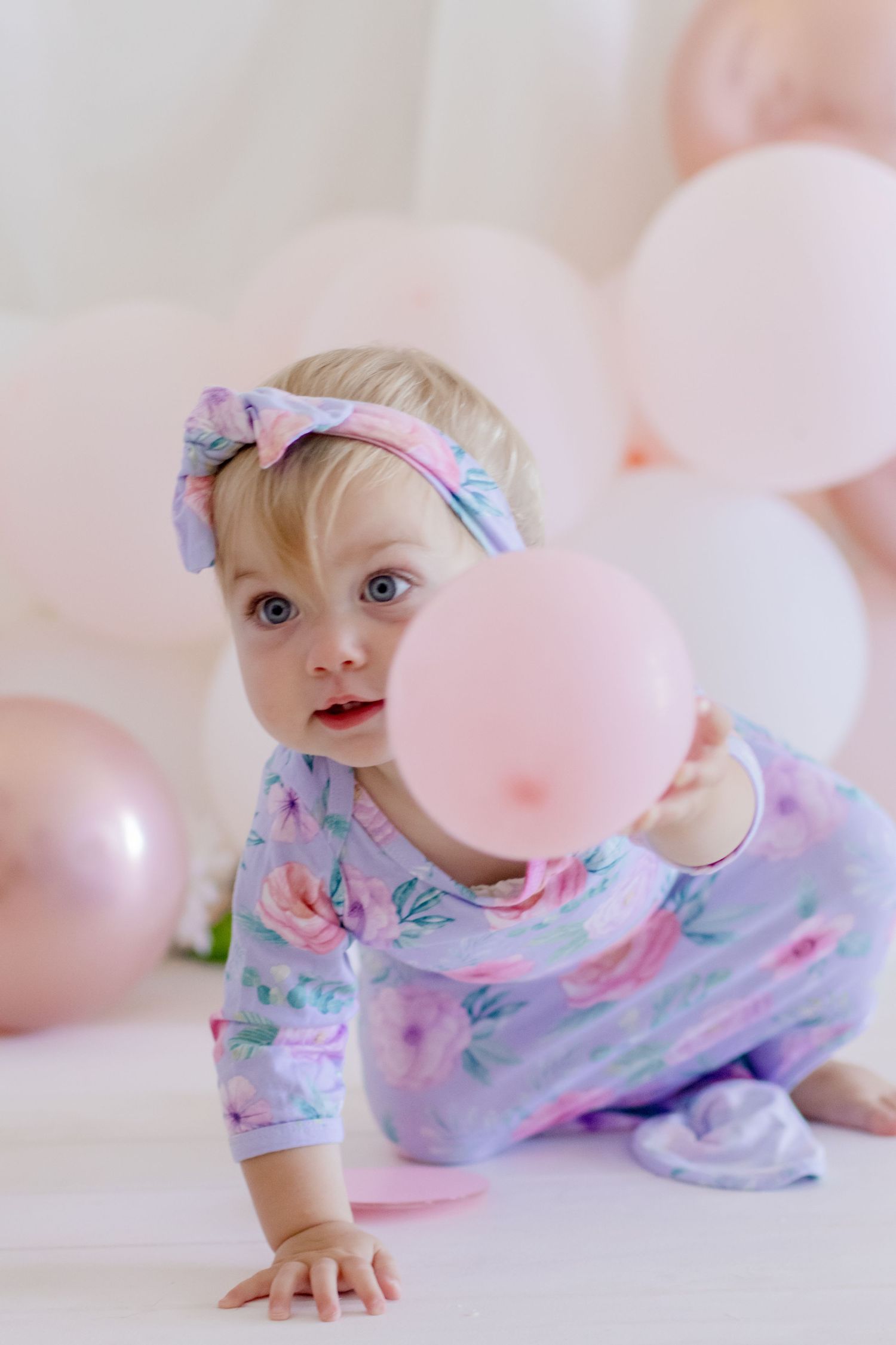 Baby in floral sleepwear reaching for pink balloons against pastel backdrop.