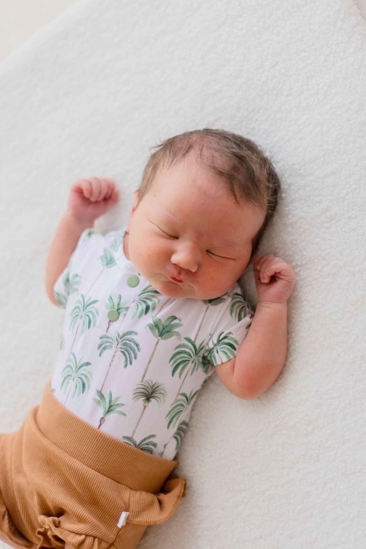 Sleeping newborn wearing tropical palm print onesie and brown shorts lying on white blanket.