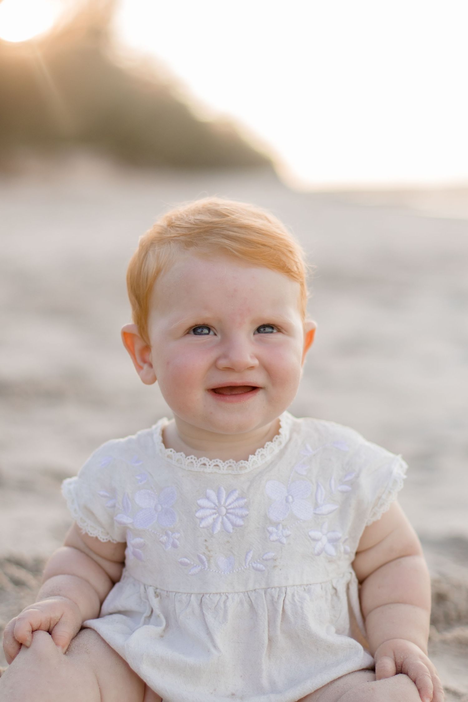 Sweet baby wearing white floral dress smiles warmly during golden hour sunset photoshoot.
