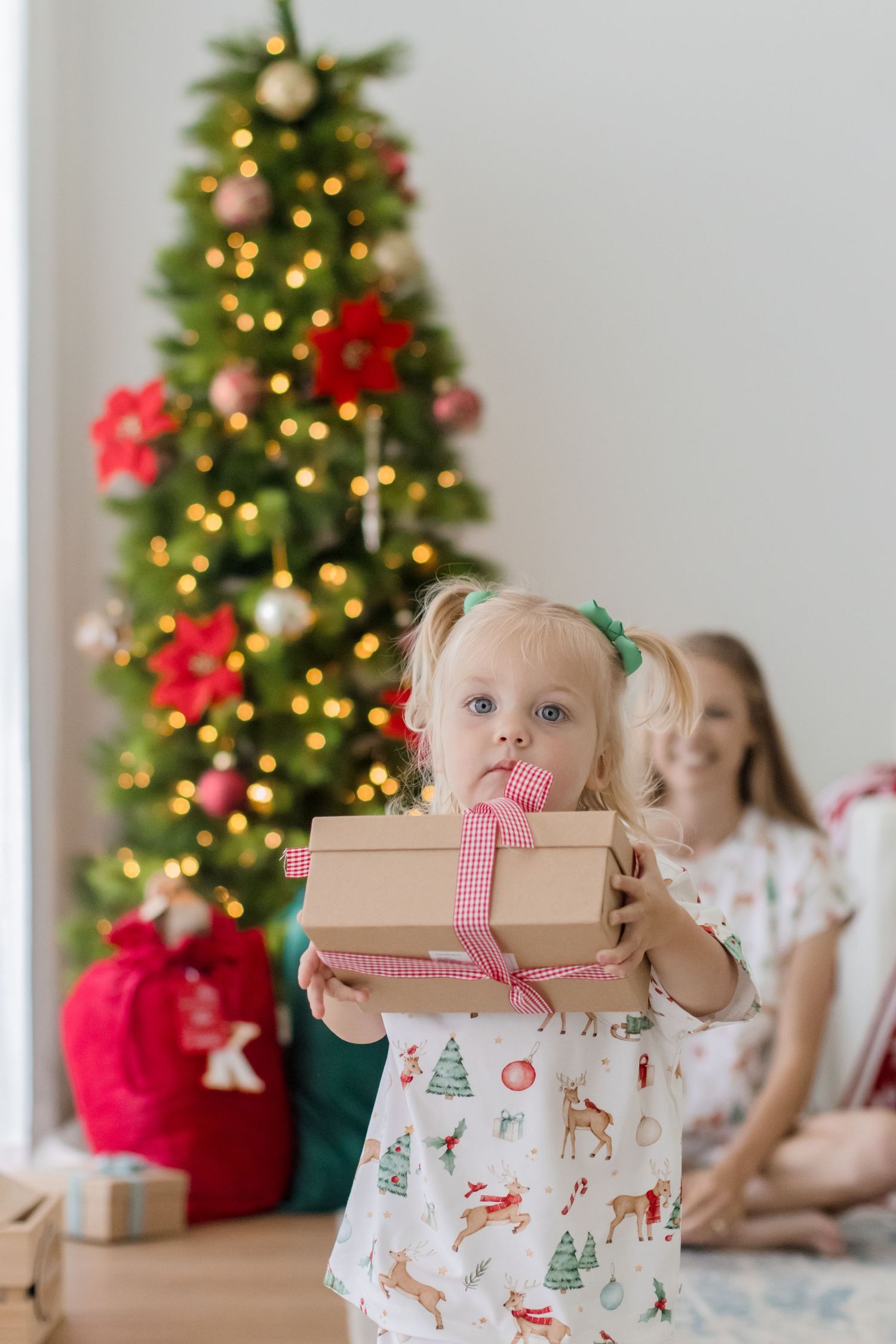 Children sit near a decorated Christmas tree with wrapped presents and holiday decor in a bright room.