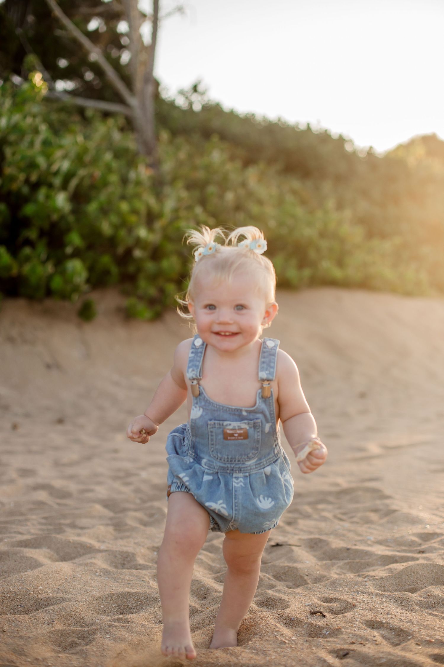 Smiling toddler in denim overalls playing on sandy beach at sunset.