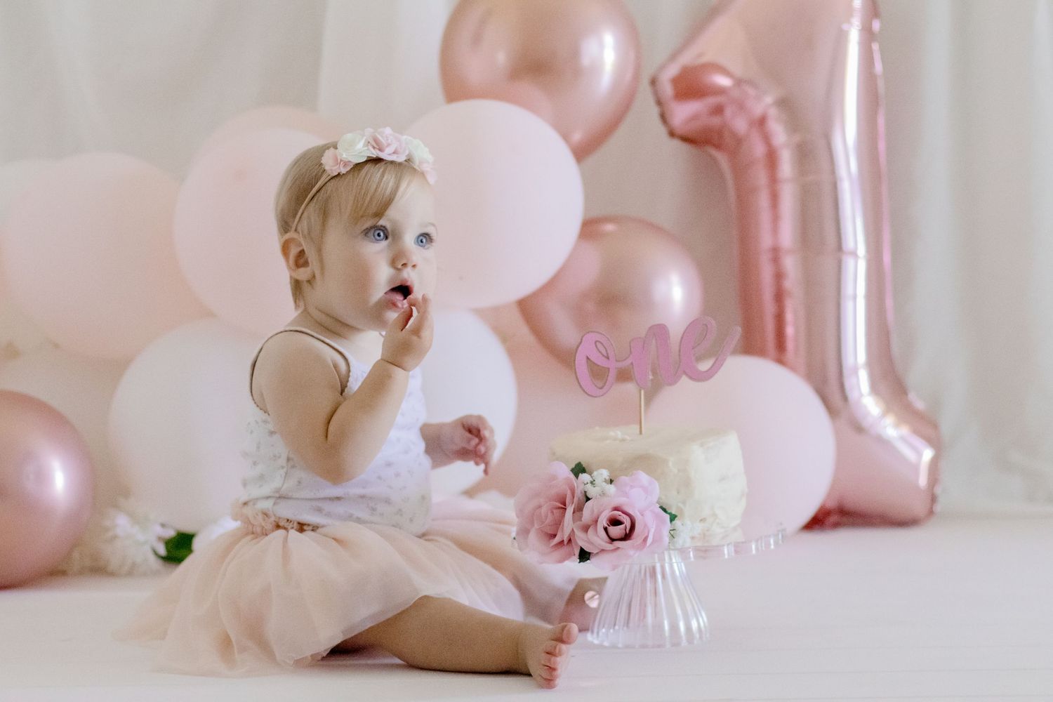 Baby exploring birthday decorations with pink balloons and number one balloon in background.