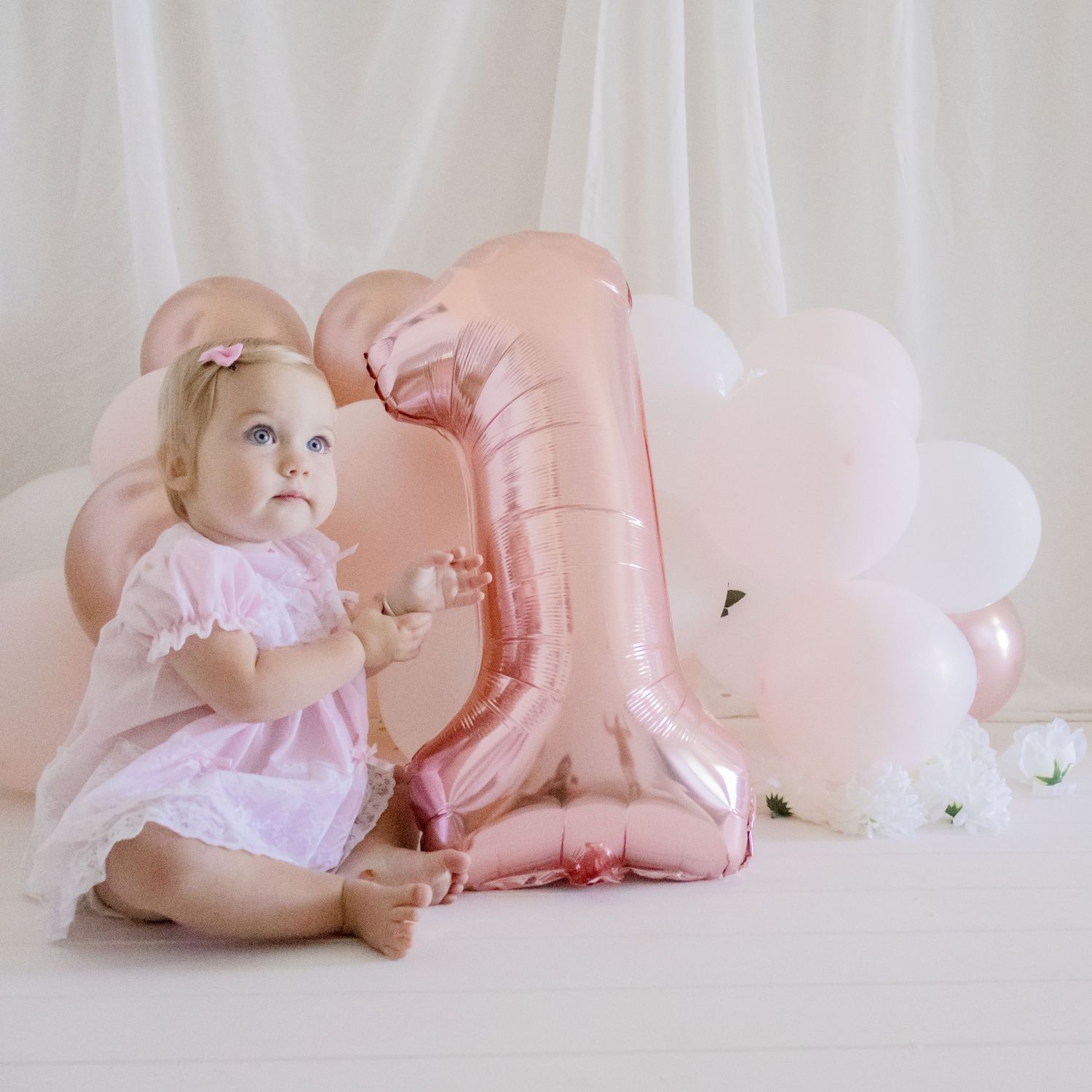 Baby in pink dress sitting beside large rose gold number one balloon and white balloons.