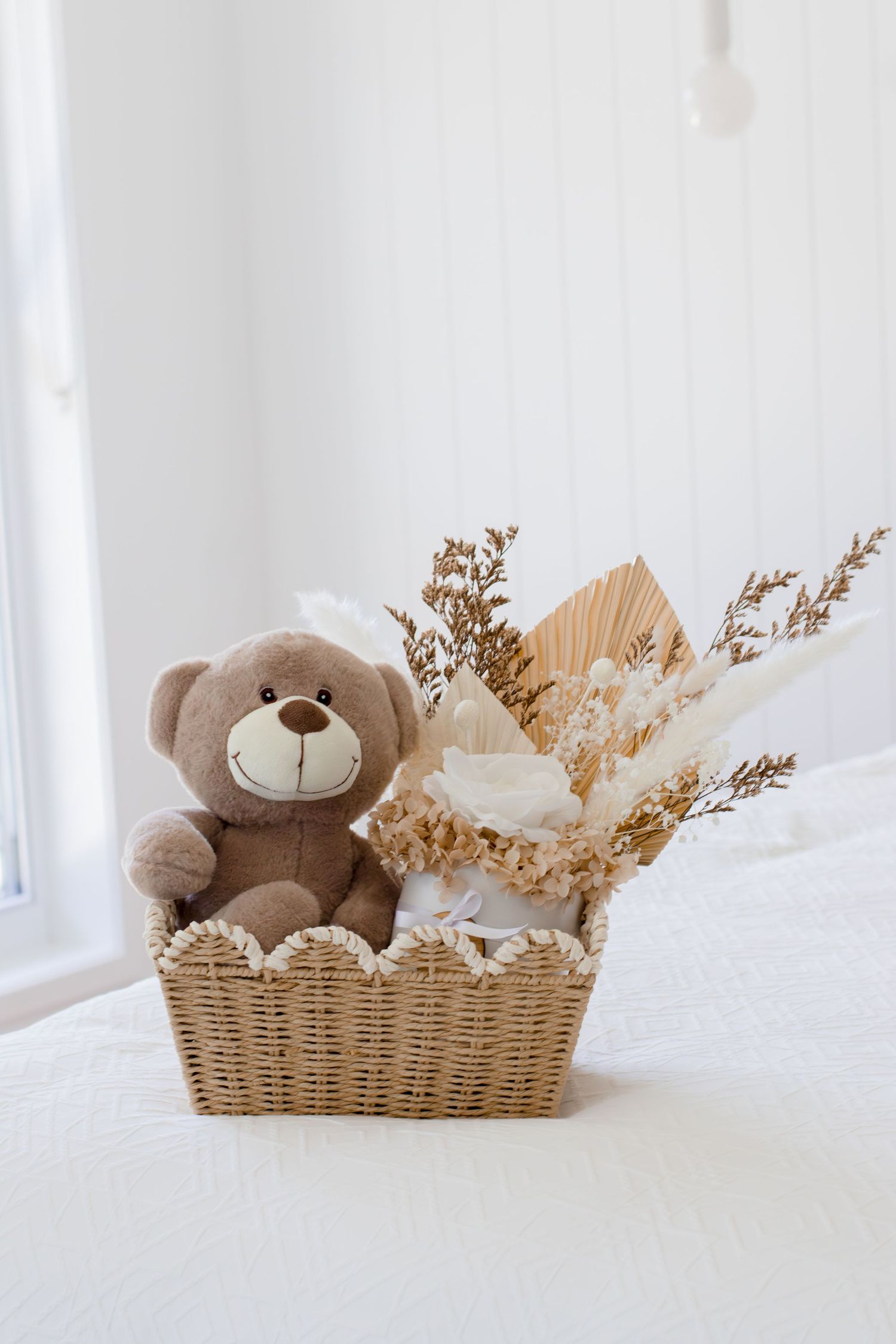 Brown teddy bear and dried floral bouquet in a wicker basket on a white bed.