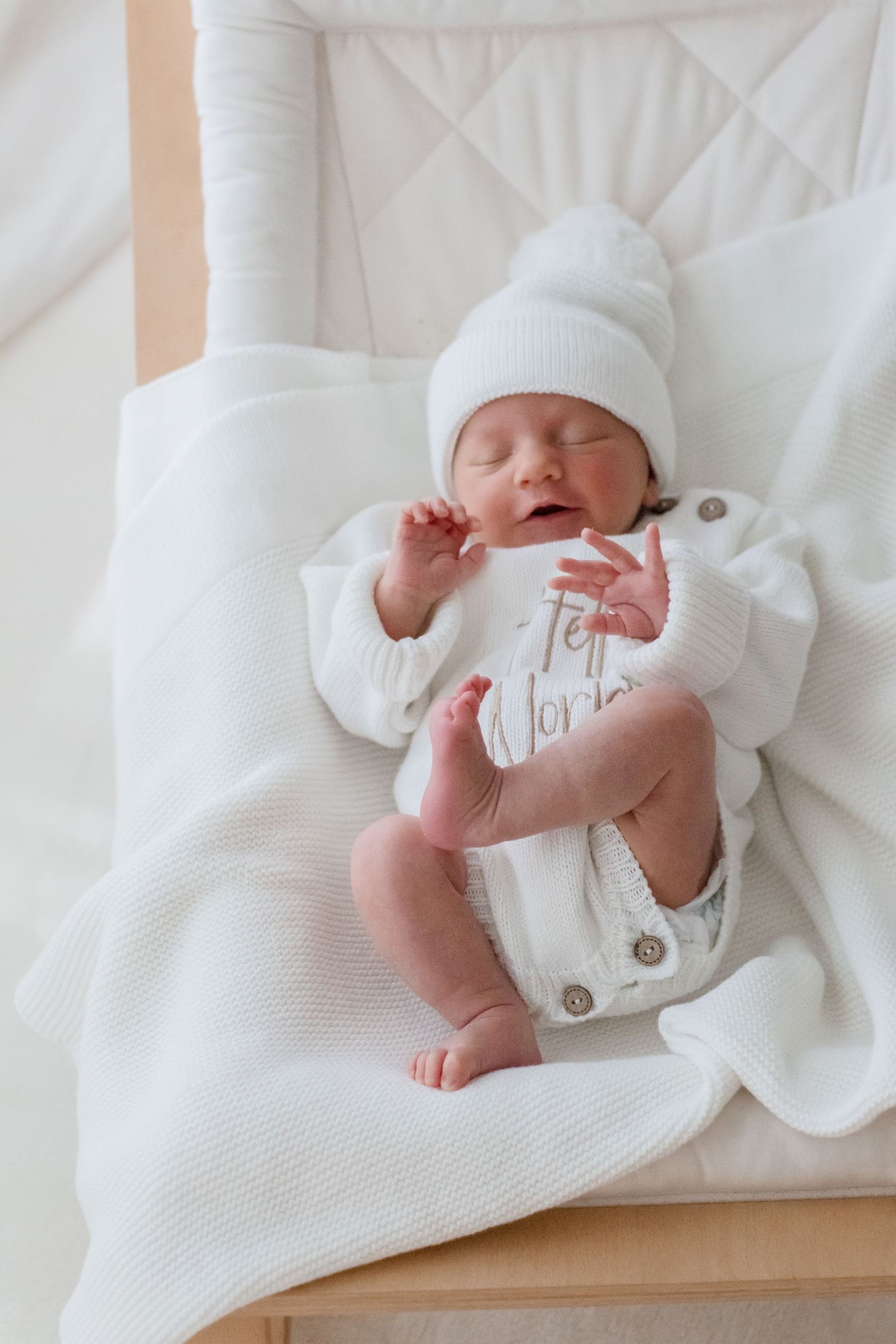 A peacefully sleeping newborn in white knit clothes and hat rests on white bedding.