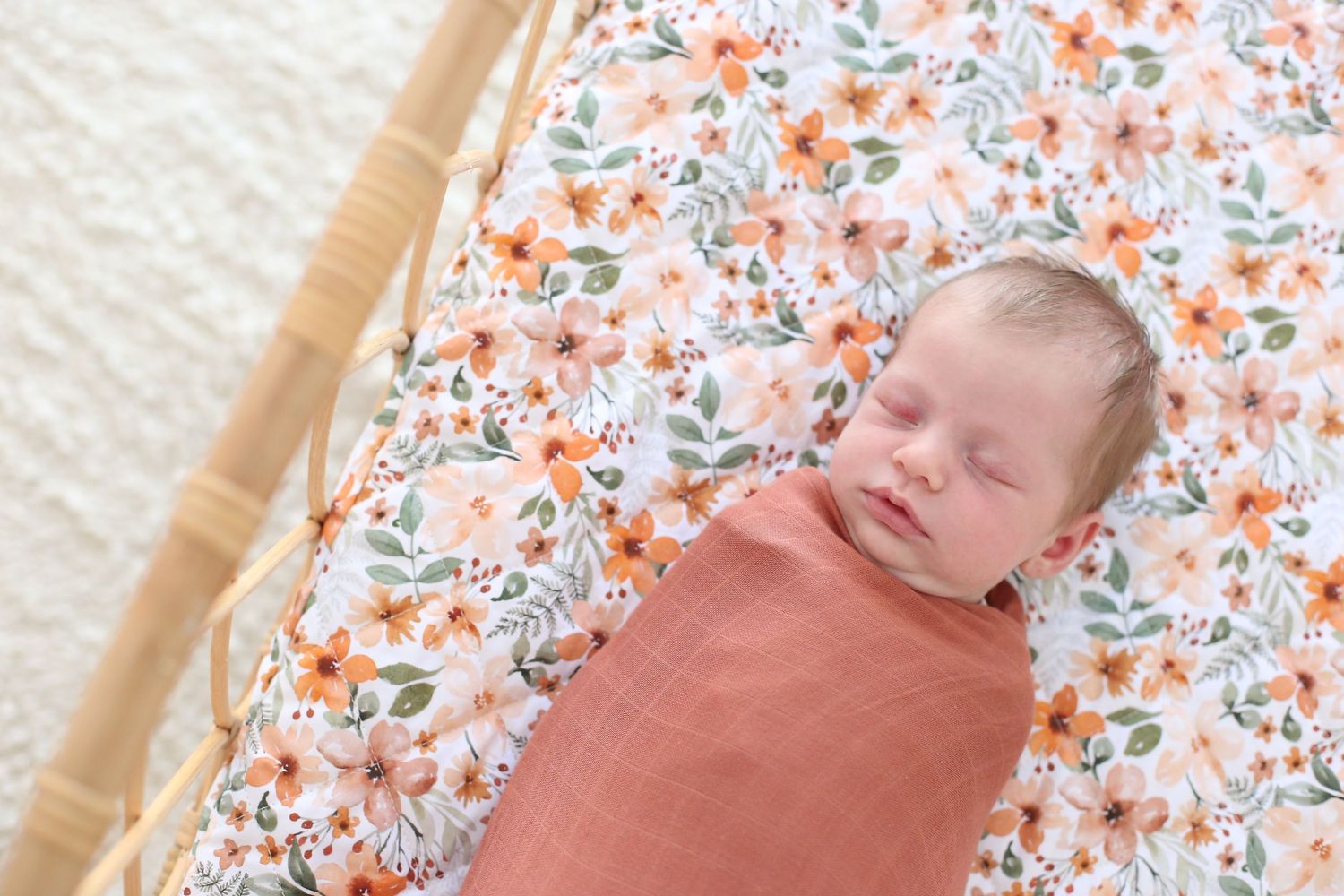 Newborn baby resting peacefully in coral colored swaddle on floral bedding.