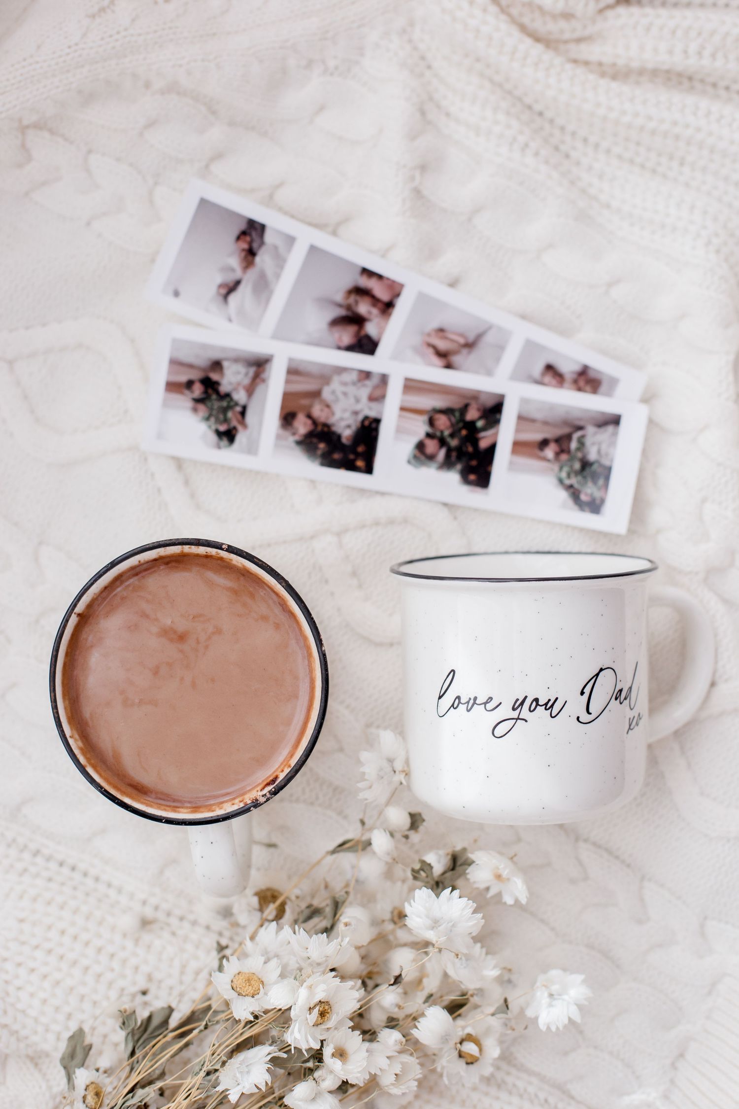 Two mugs of hot chocolate with photos and white dried flowers on a textured white surface.