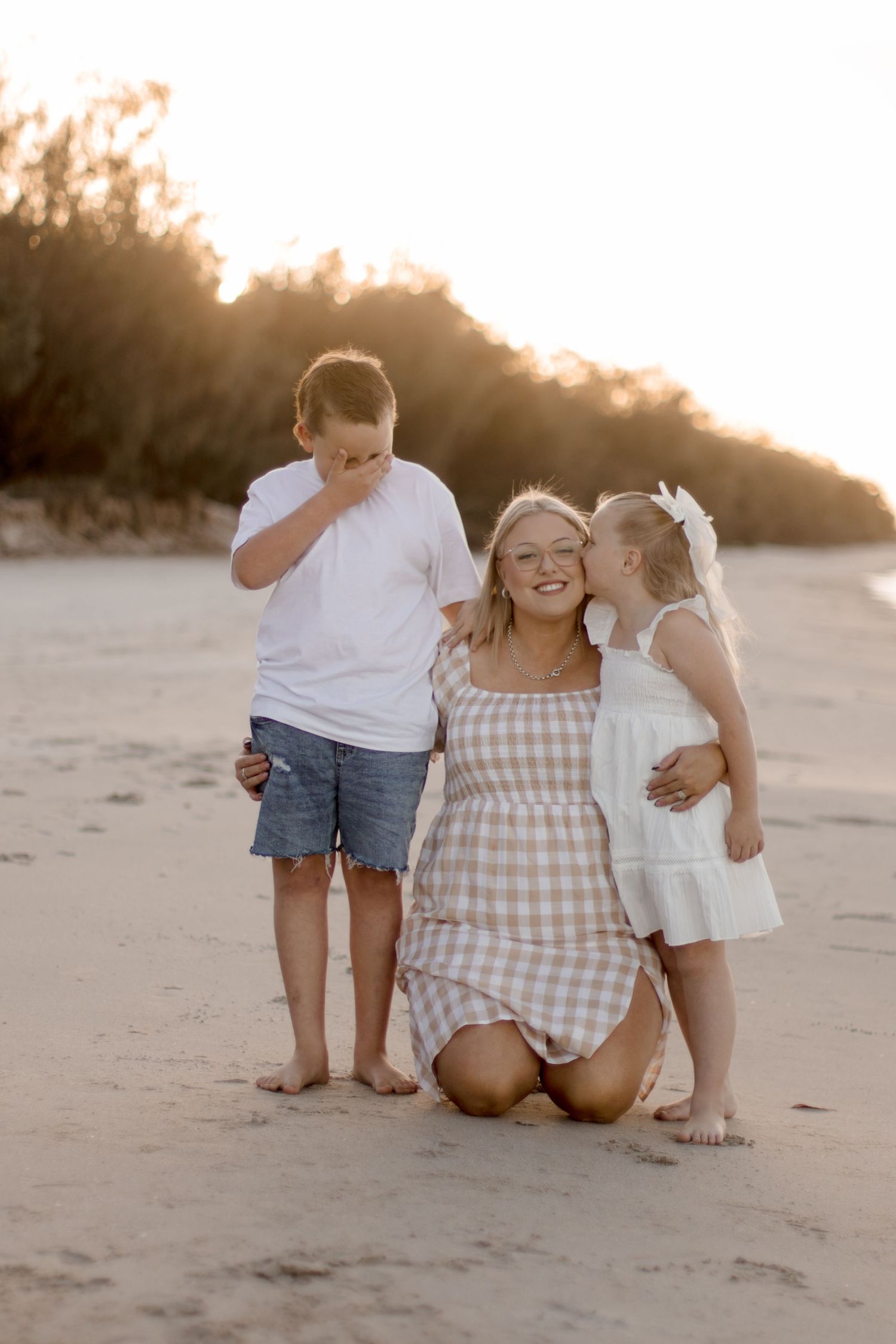 A family enjoys a candid moment together on the beach at sunset, laughing and smiling in casual summer attire.