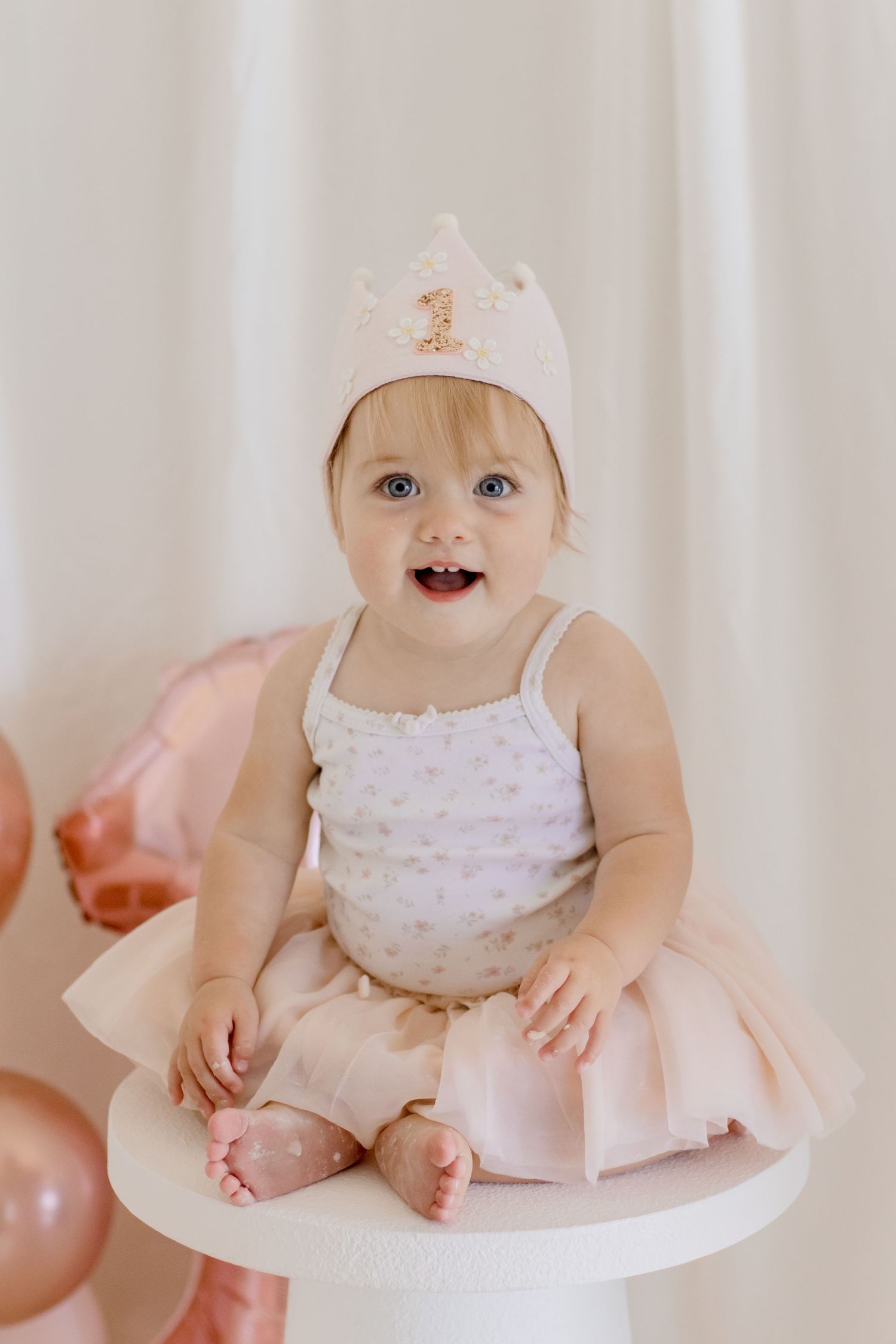Smiling baby in crown and pink dress sits on white pedestal during birthday celebration.