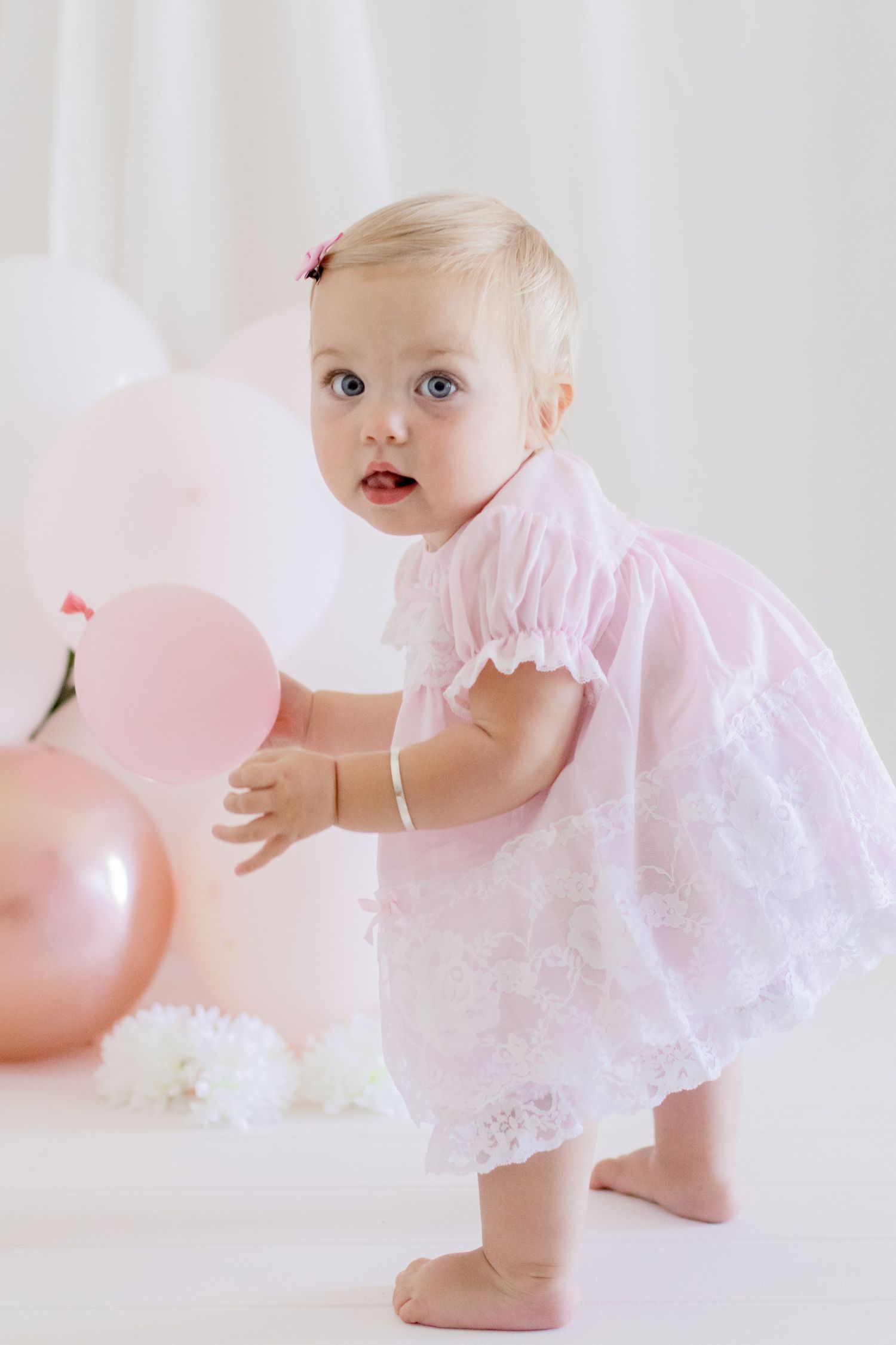 Baby in white lace dress reaching for pink balloons against white backdrop.