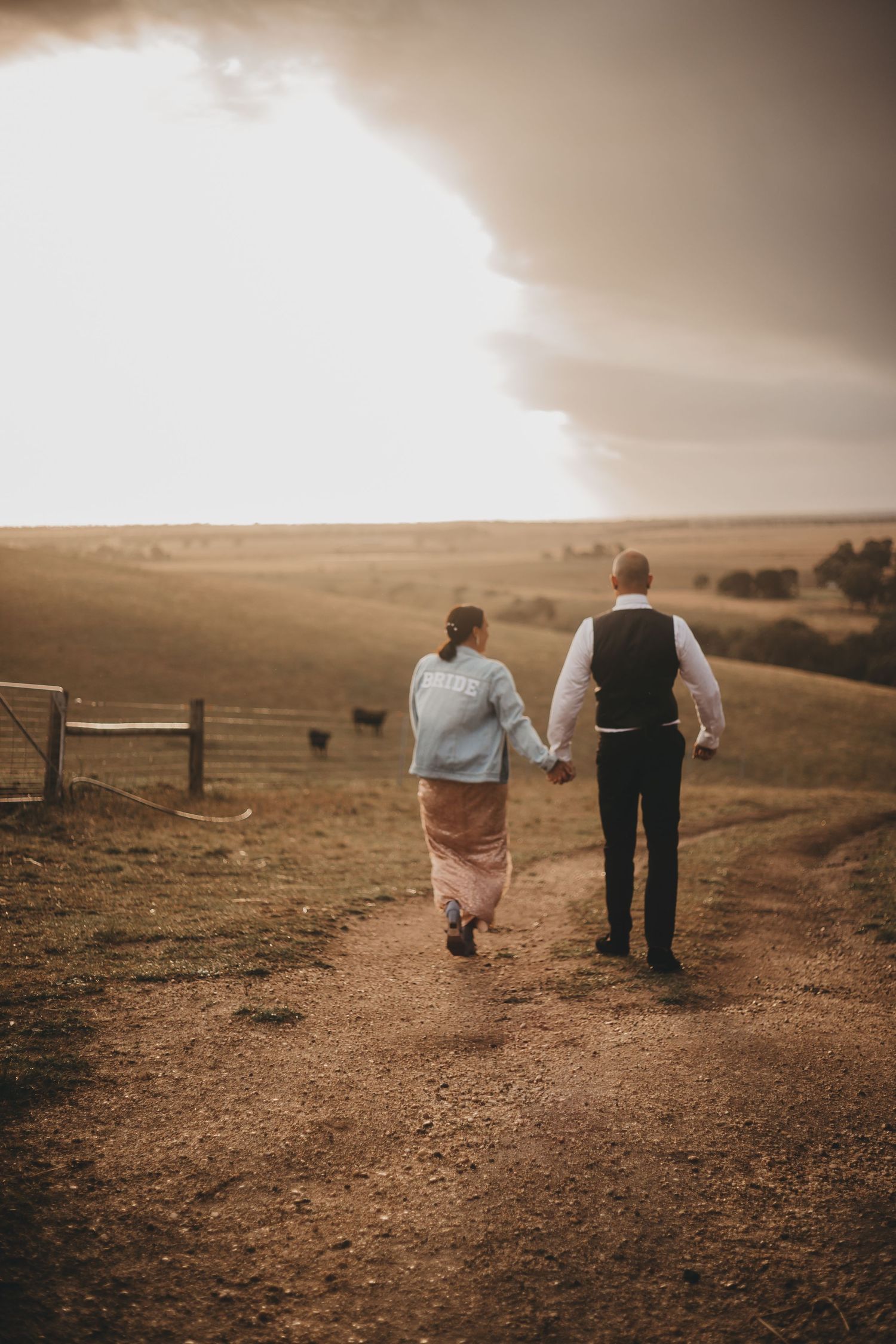 Couple walking hand in hand on a dirt path through rolling hills during golden hour sunset.