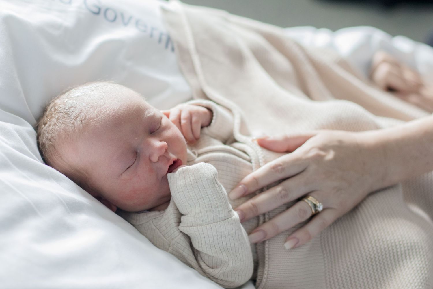 A sleeping newborn in soft knitwear being gently touched by a hand with a wedding ring.