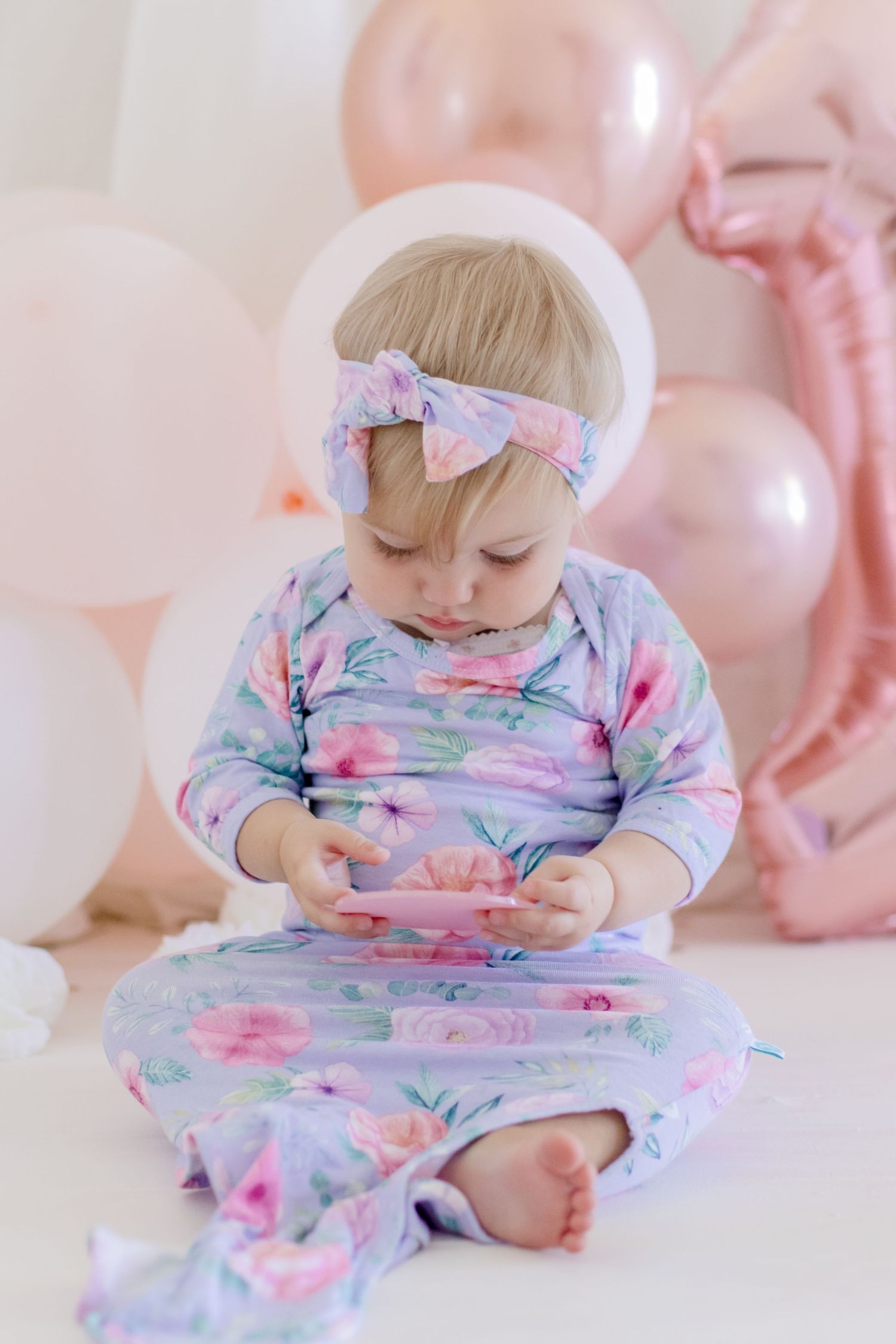 A floral onesie outfit and bow headband amid pink balloons in a dreamy photoshoot setting.