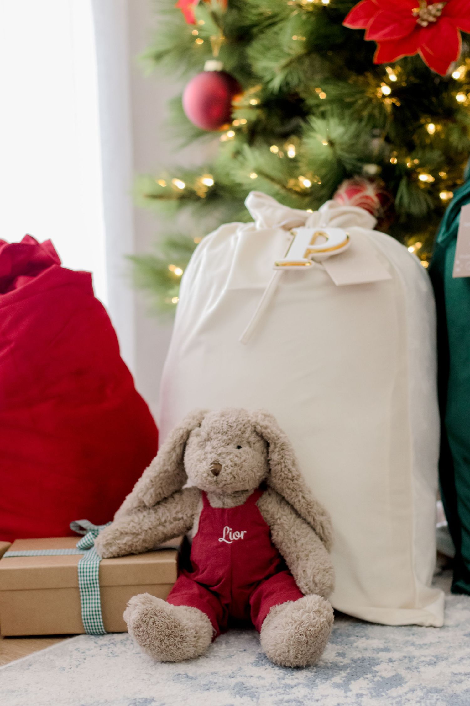 A plush gray bunny in red overalls sits beside Christmas presents and gift bags under a decorated tree with twinkling lights.