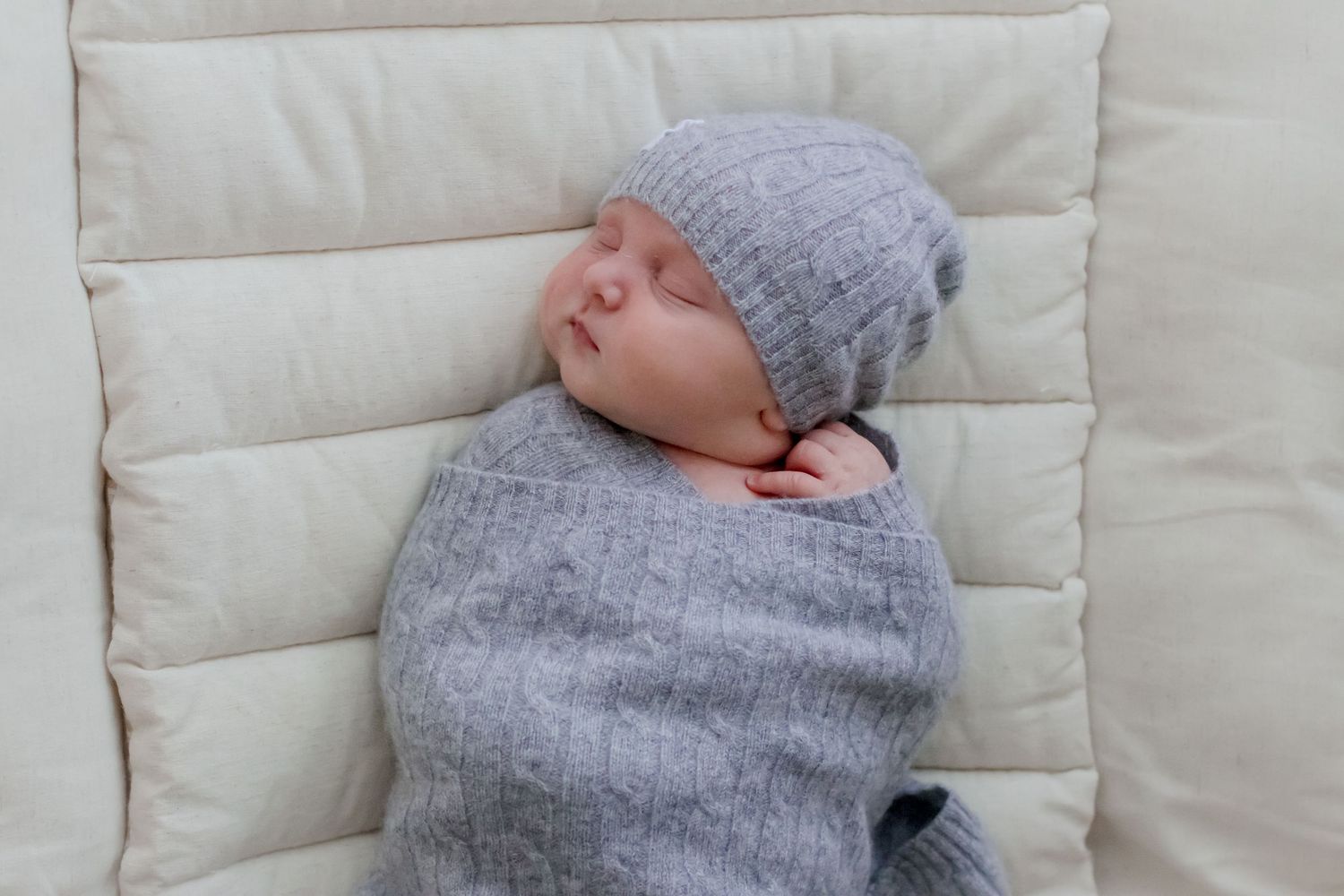 Peaceful sleeping newborn wearing a cozy gray knit sweater and matching beanie on white cushioned surface.