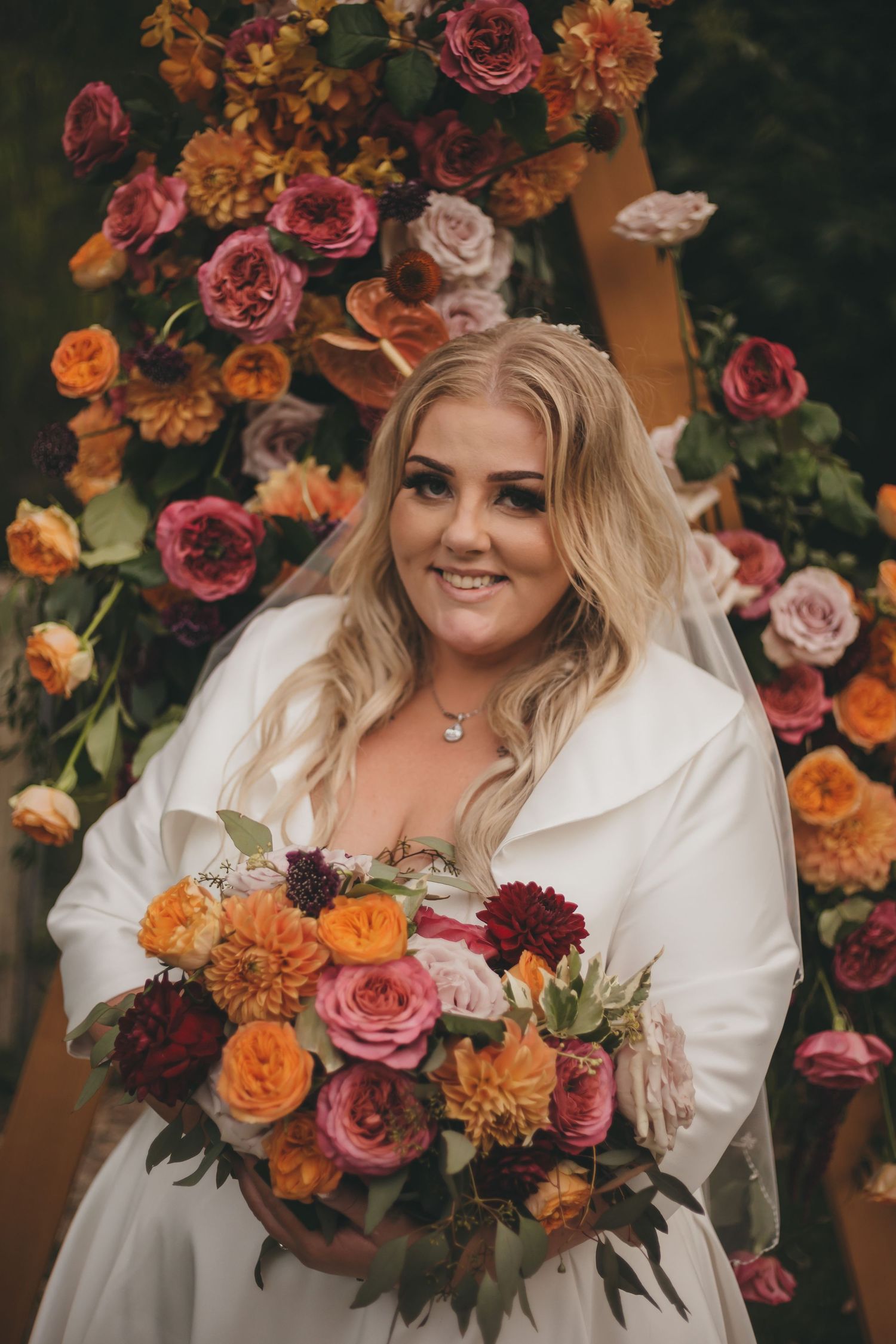 Beautiful autumn wedding photo with bride holding vibrant bouquet of orange, pink and burgundy roses against floral arch.