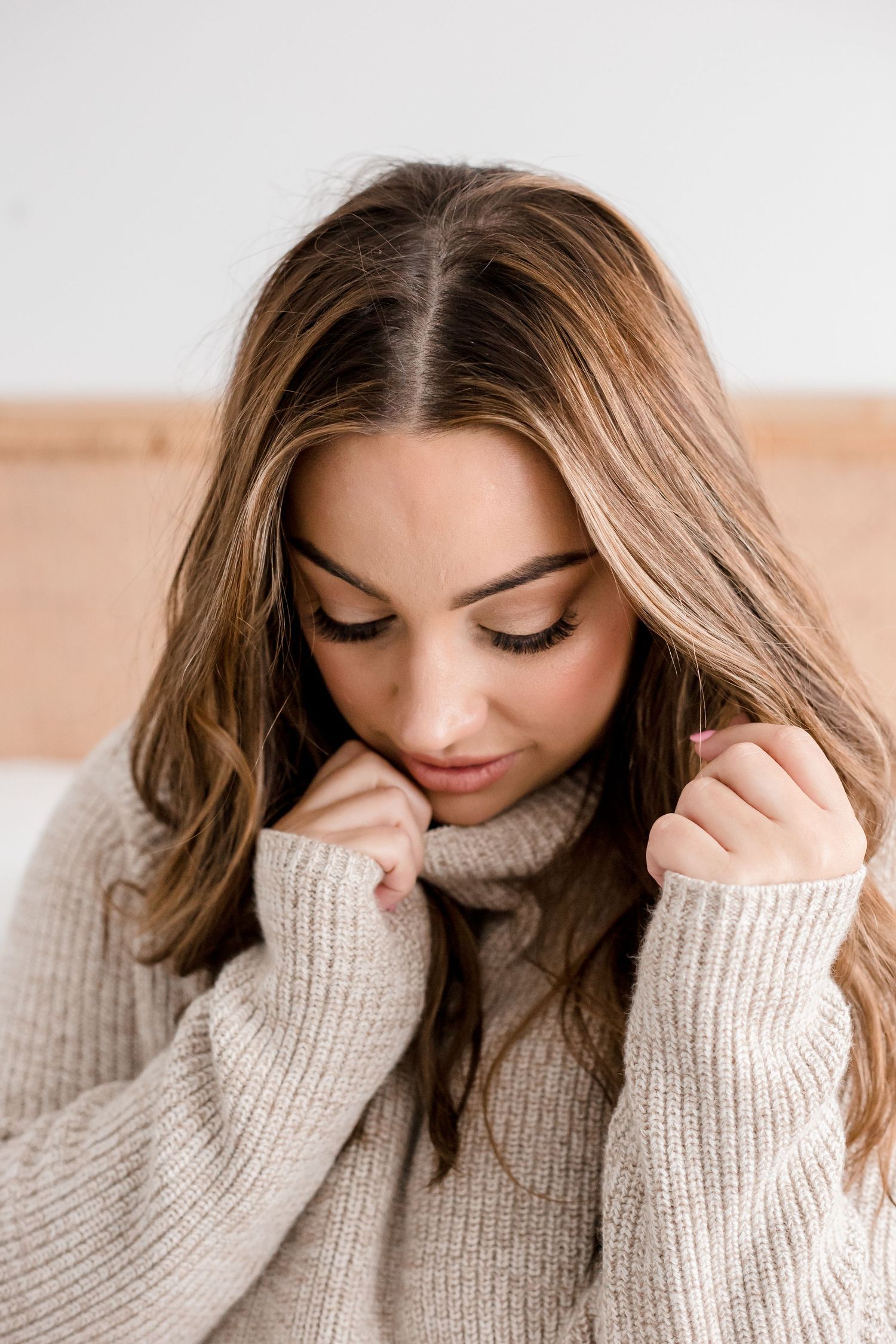 Woman in beige knitted sweater poses in soft lighting with natural makeup and downward gaze.