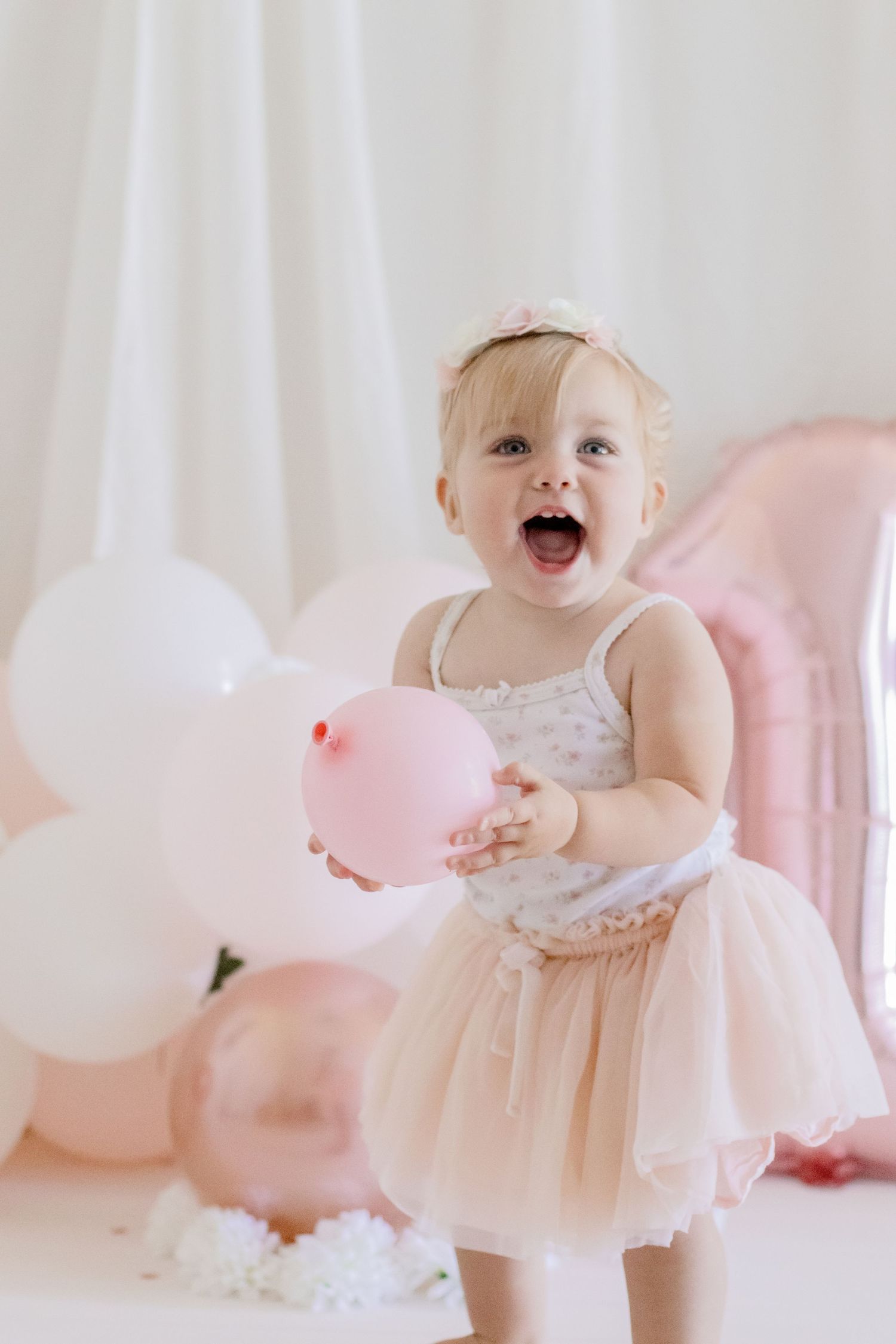 Happy toddler laughing while playing with pink and rose gold balloons in tulle dress.