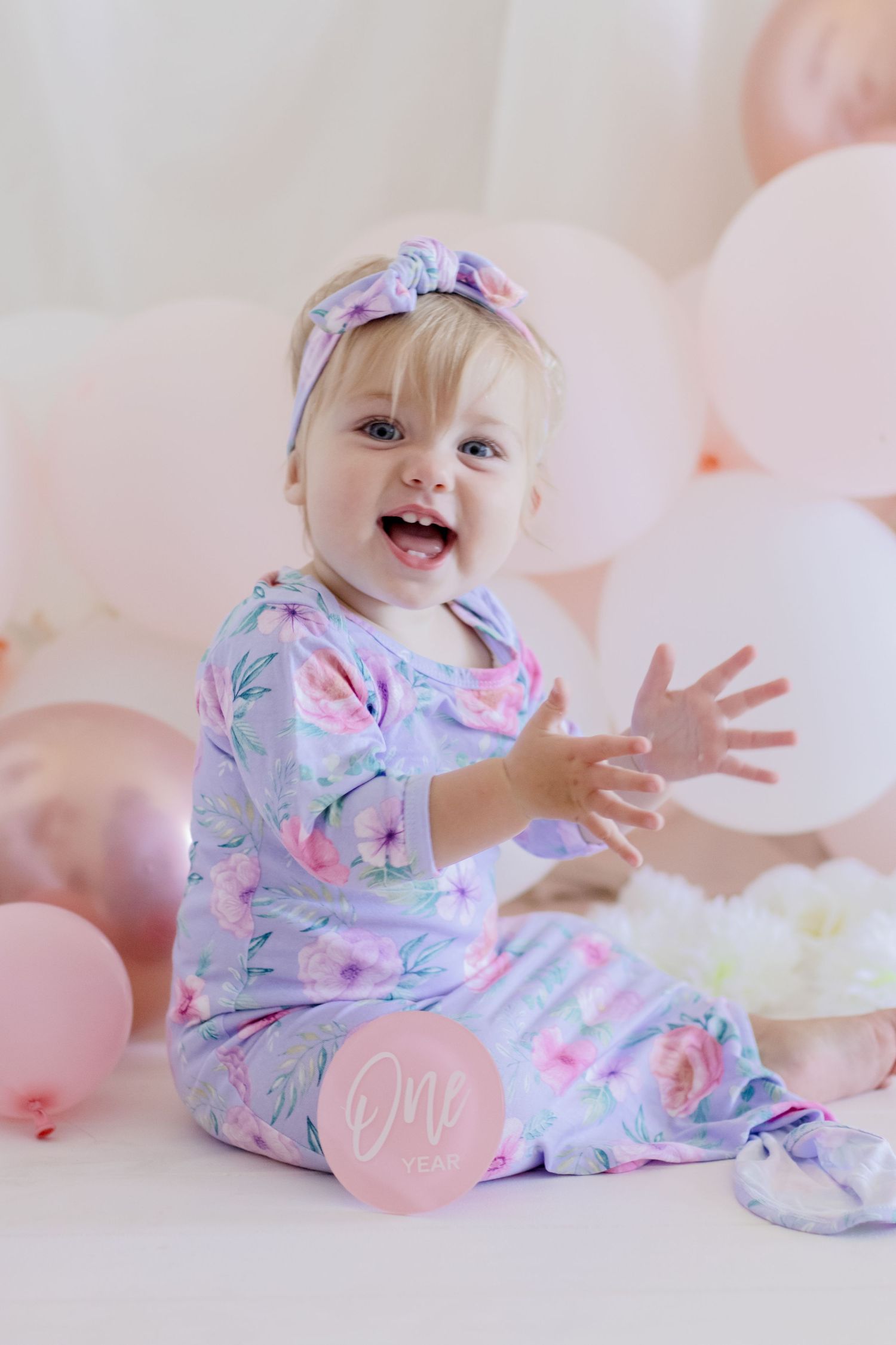 Playful infant in floral pajamas surrounded by pink and white balloons.