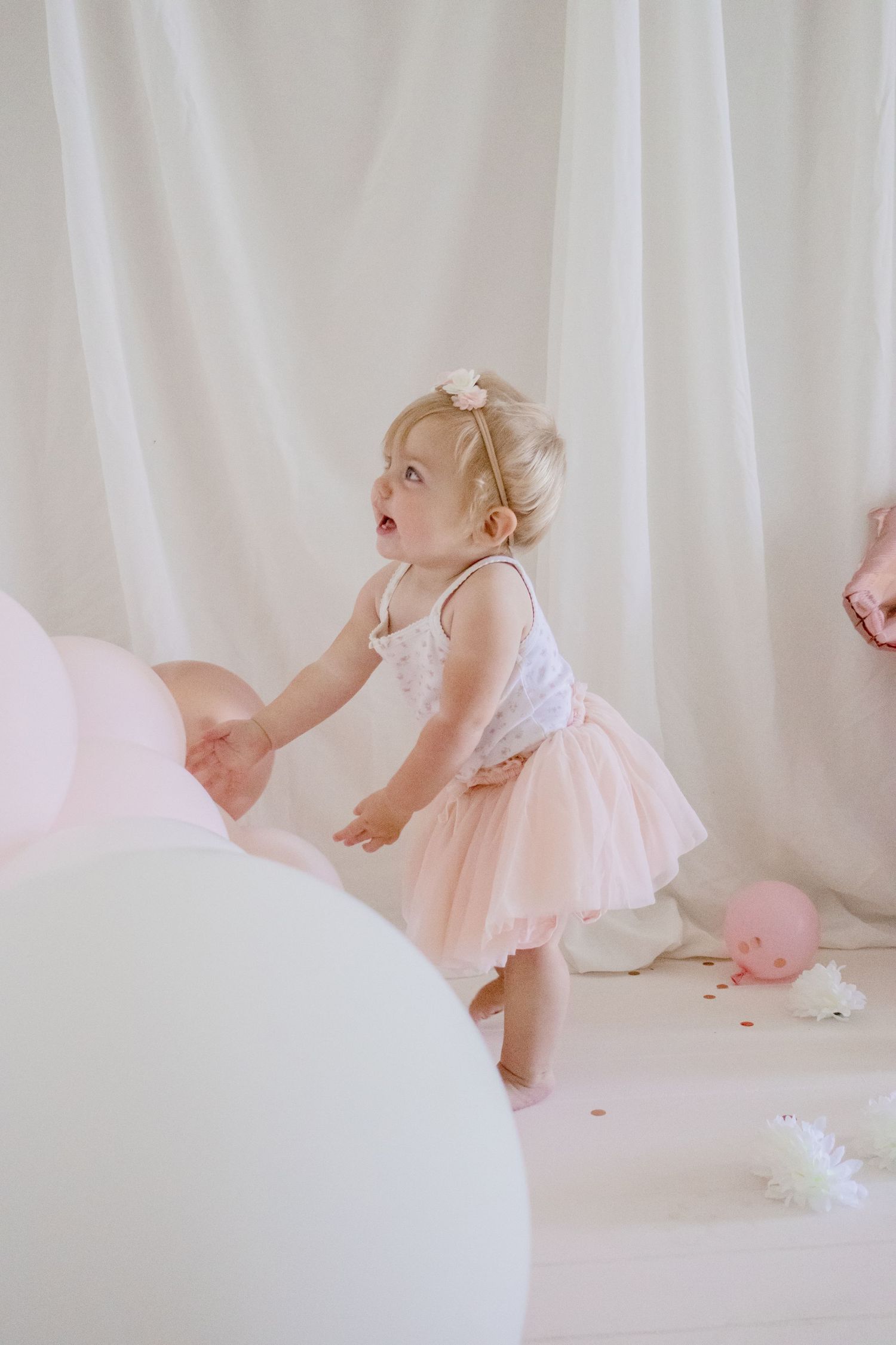 A small child in a pink tulle dress reaches for white balloons during a festive photo session.