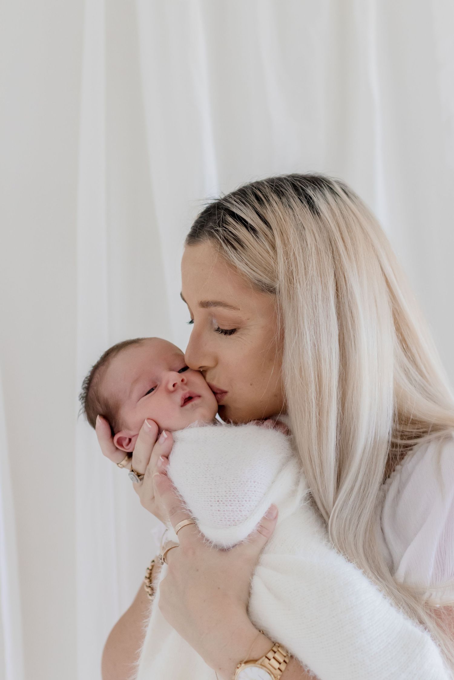 Tender moment between mother and swaddled newborn baby against white curtain background.