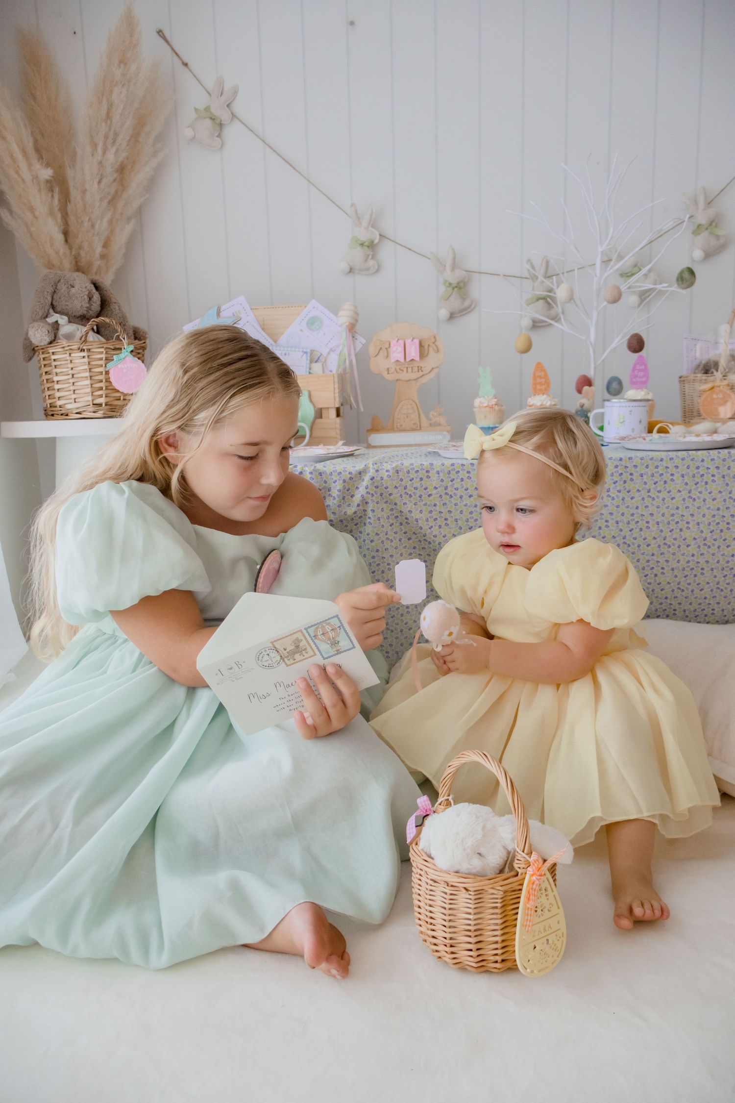 Two children in pastel dresses sharing storytime with Easter decorations in background.