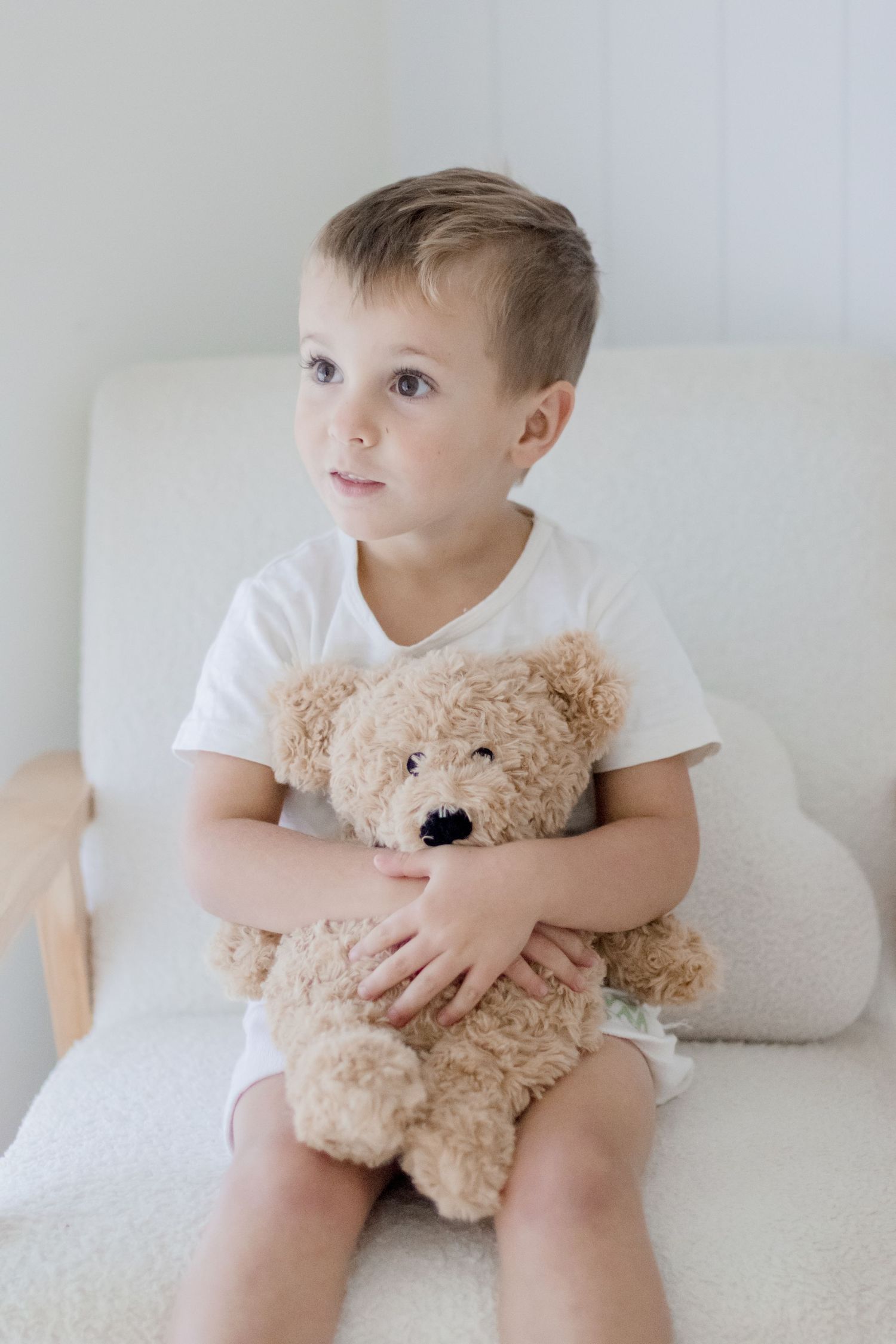Young child in white shirt holding brown teddy bear while sitting on white chair.