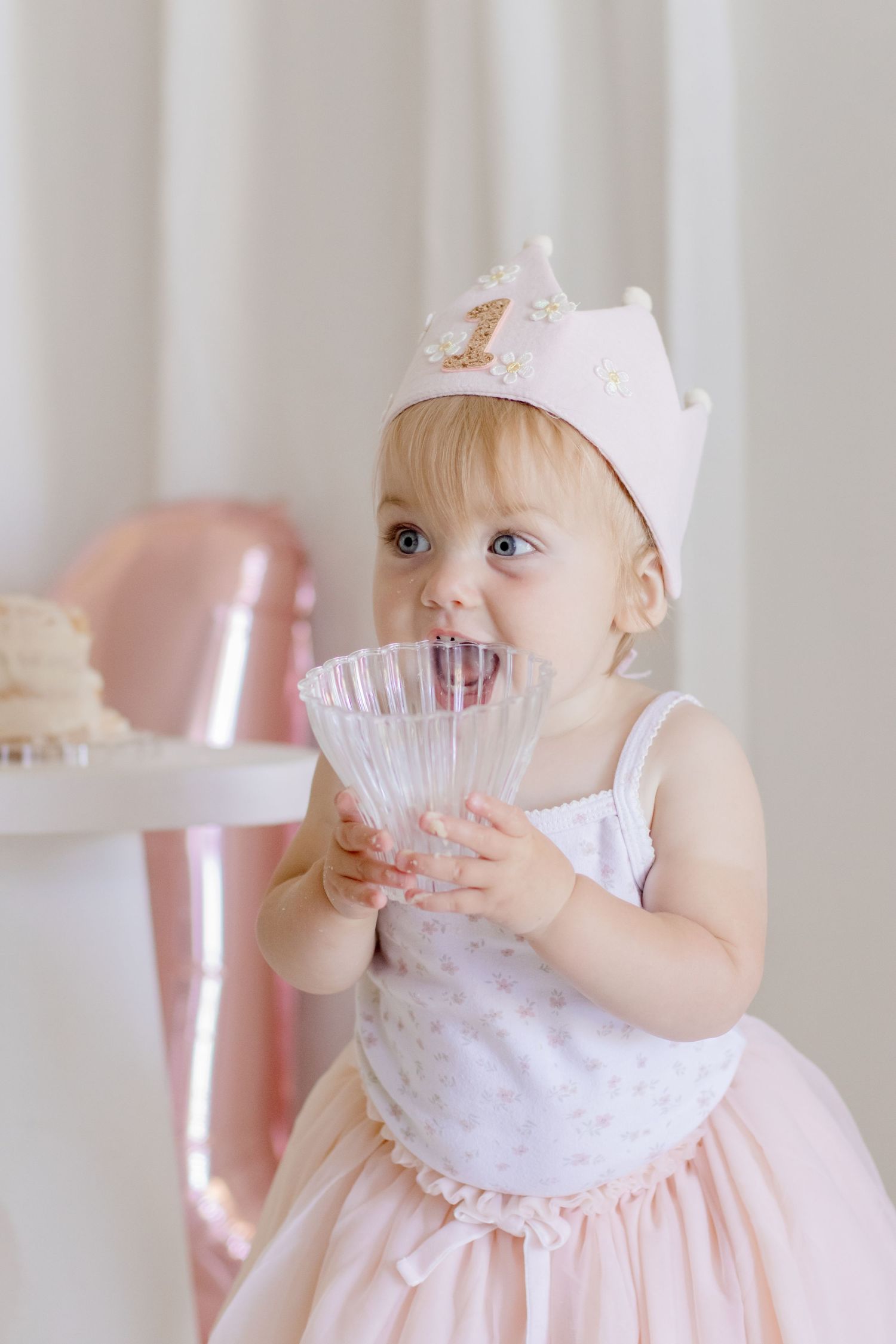 Baby wearing crown and pink dress holds crystal glass during birthday celebration.