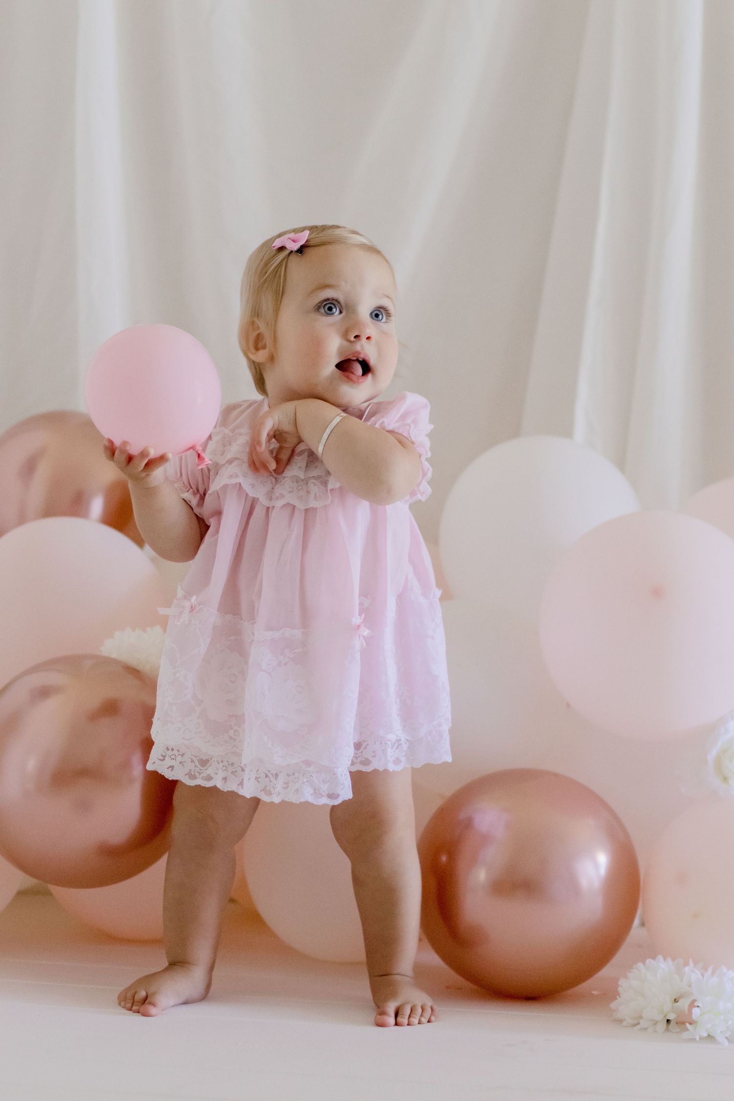 Baby in pink dress reaches up while standing with balloons.