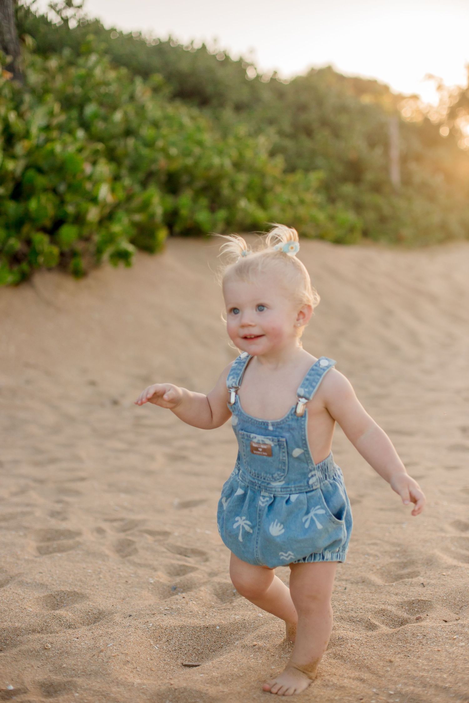A toddler in blue overalls plays joyfully on a sandy beach at sunset with greenery in the background.