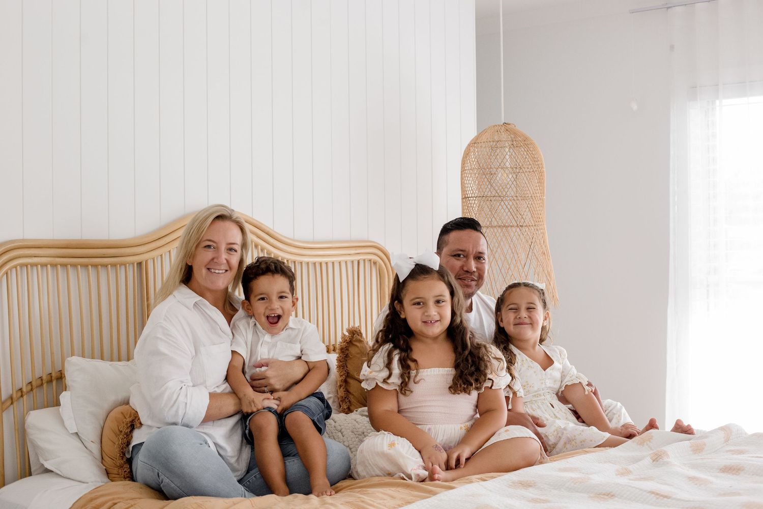 Family poses together on beige bed with rattan headboard in bright white room.