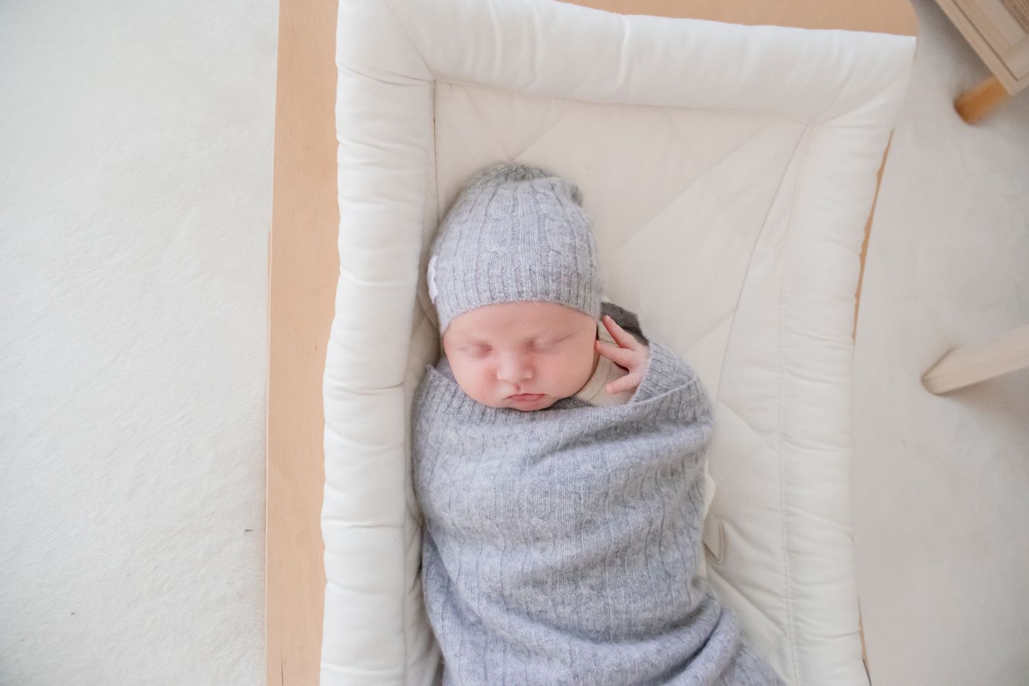 Newborn baby sleeping peacefully while swaddled in soft gray blanket with matching knit cap on white bedding.