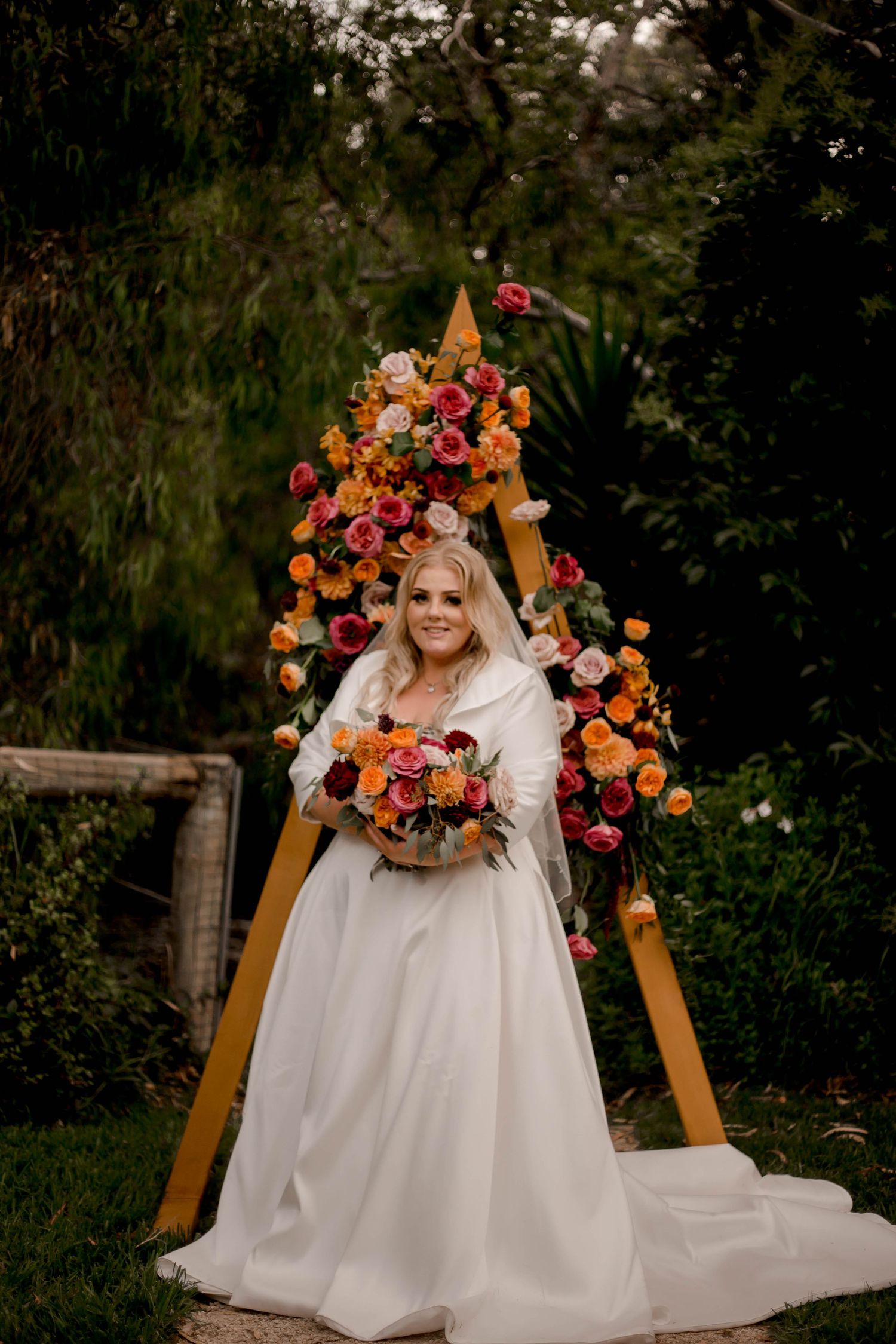A bride in a white wedding dress poses beneath a wooden arch adorned with colorful autumn roses and florals in a garden.