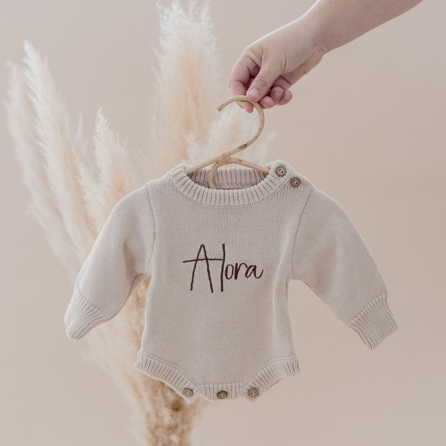 Hand holds a small beige baby onesie with 'Adora' text against neutral backdrop with pampas grass decor.