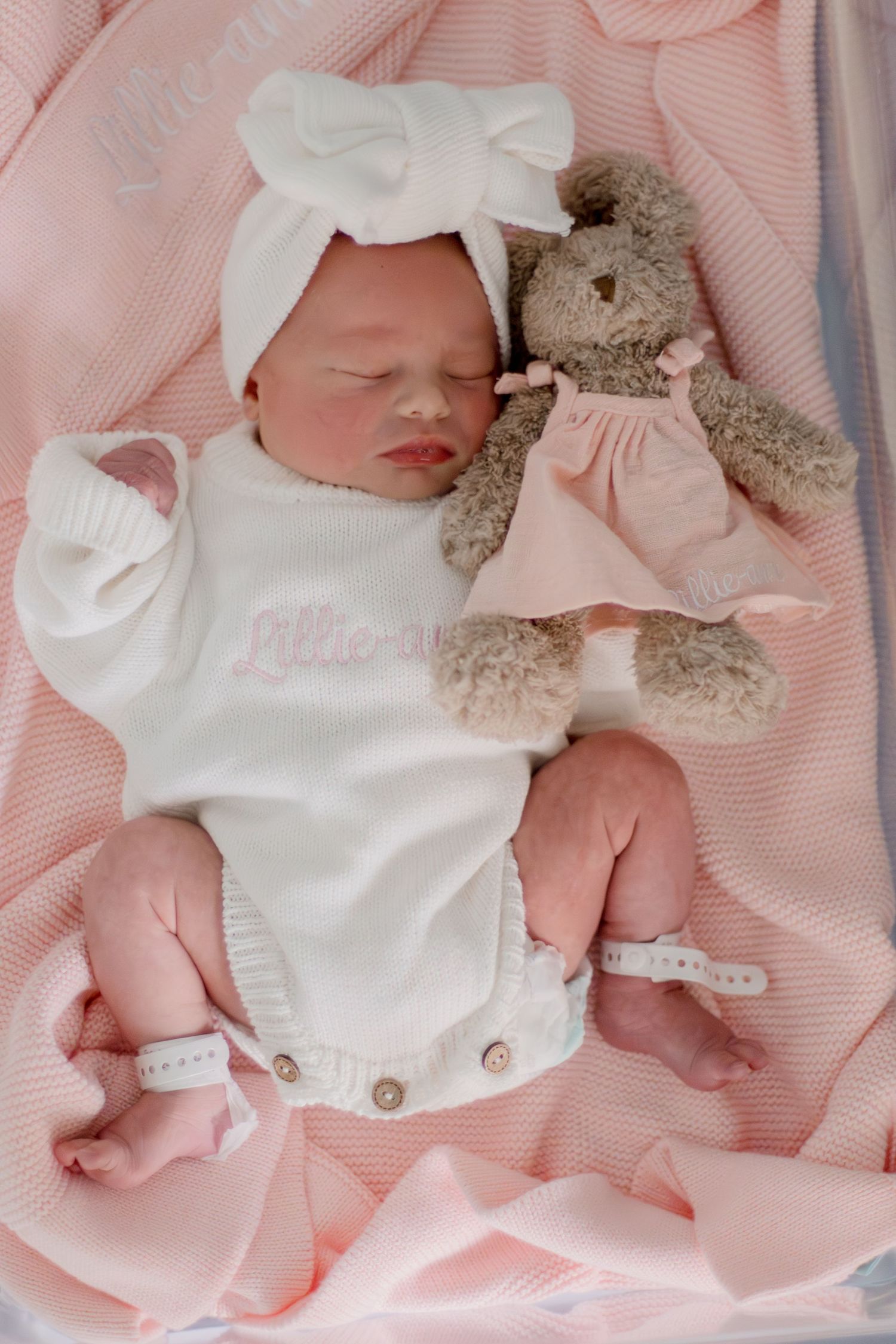 Sleeping newborn baby girl in white onesie with bow headband beside a plush bunny on pink blanket.
