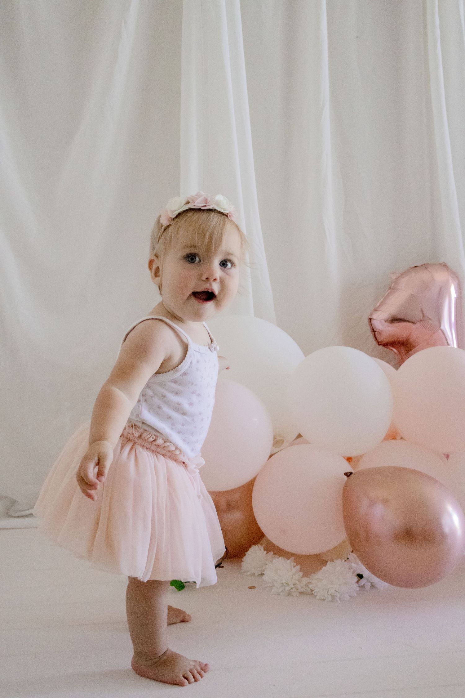 A toddler in a white and pink tulle dress stands excitedly among pink and rose gold balloons against a white backdrop.