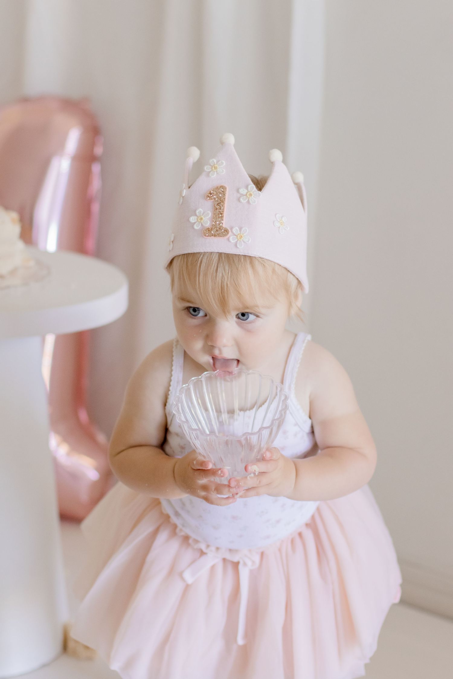 Baby in white crown and pink dress standing near birthday cake and pink balloons.