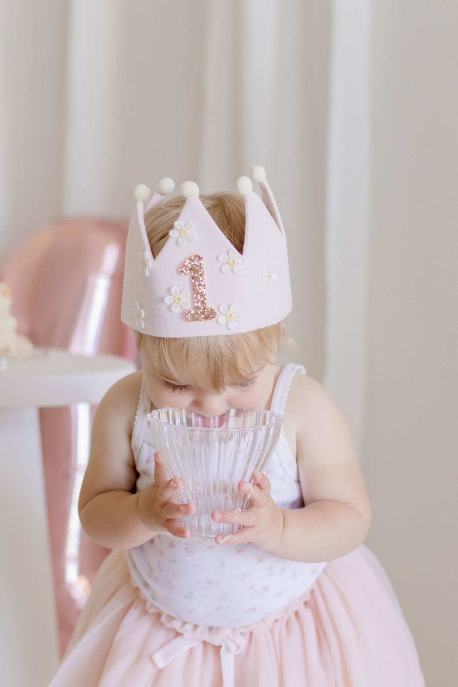 Baby wearing white crown with number 1 holds crystal glass in soft pink party setting.