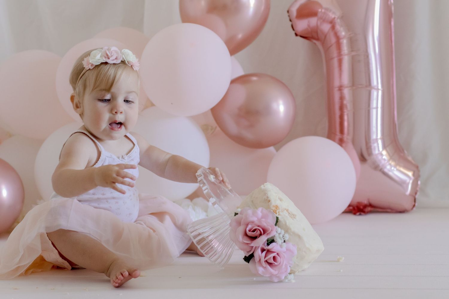 A joyful cake smash photo with pink balloons and roses against a white backdrop.