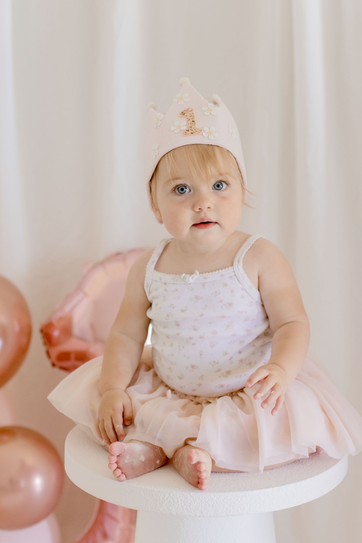 Baby in tulle dress and crown sits on white pedestal with pink balloons in background.