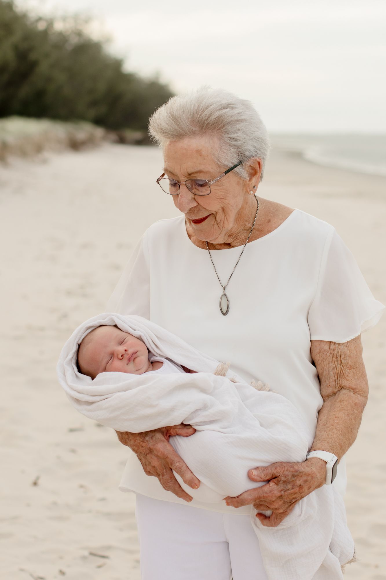 An elderly woman in white holds and gazes lovingly at a swaddled baby on a beach.