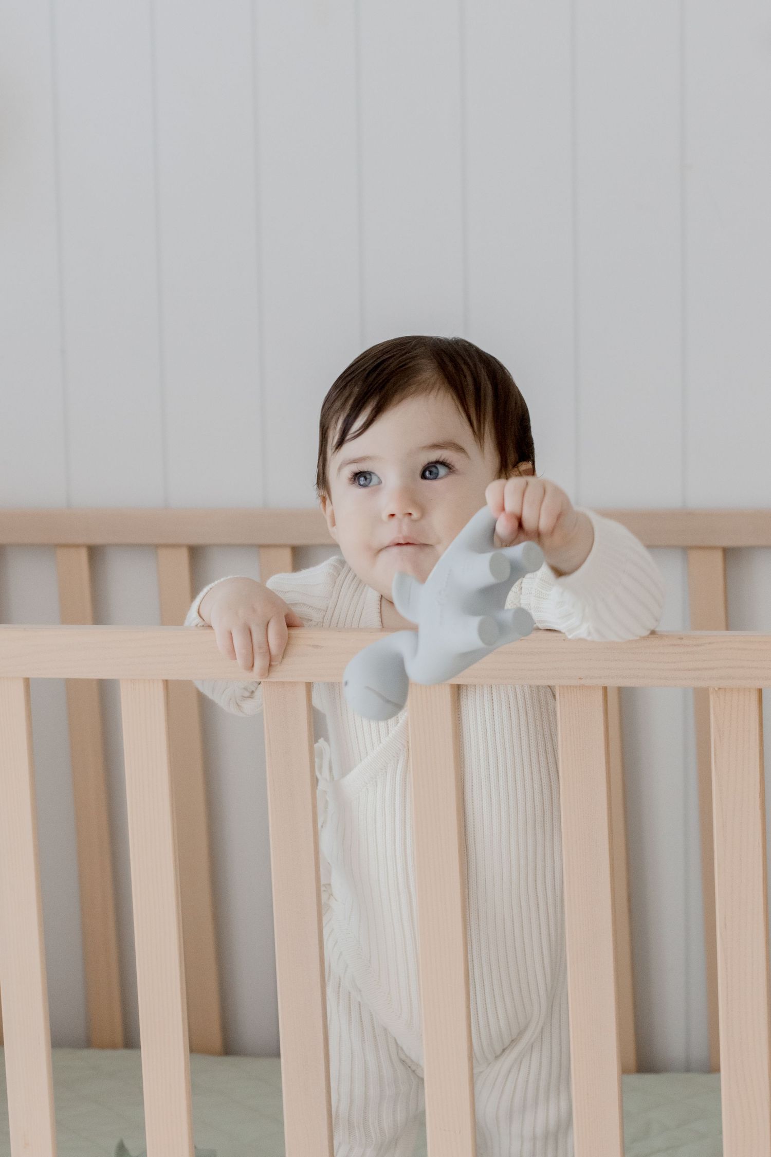 Small child in white ribbed outfit standing in wooden crib looking up.
