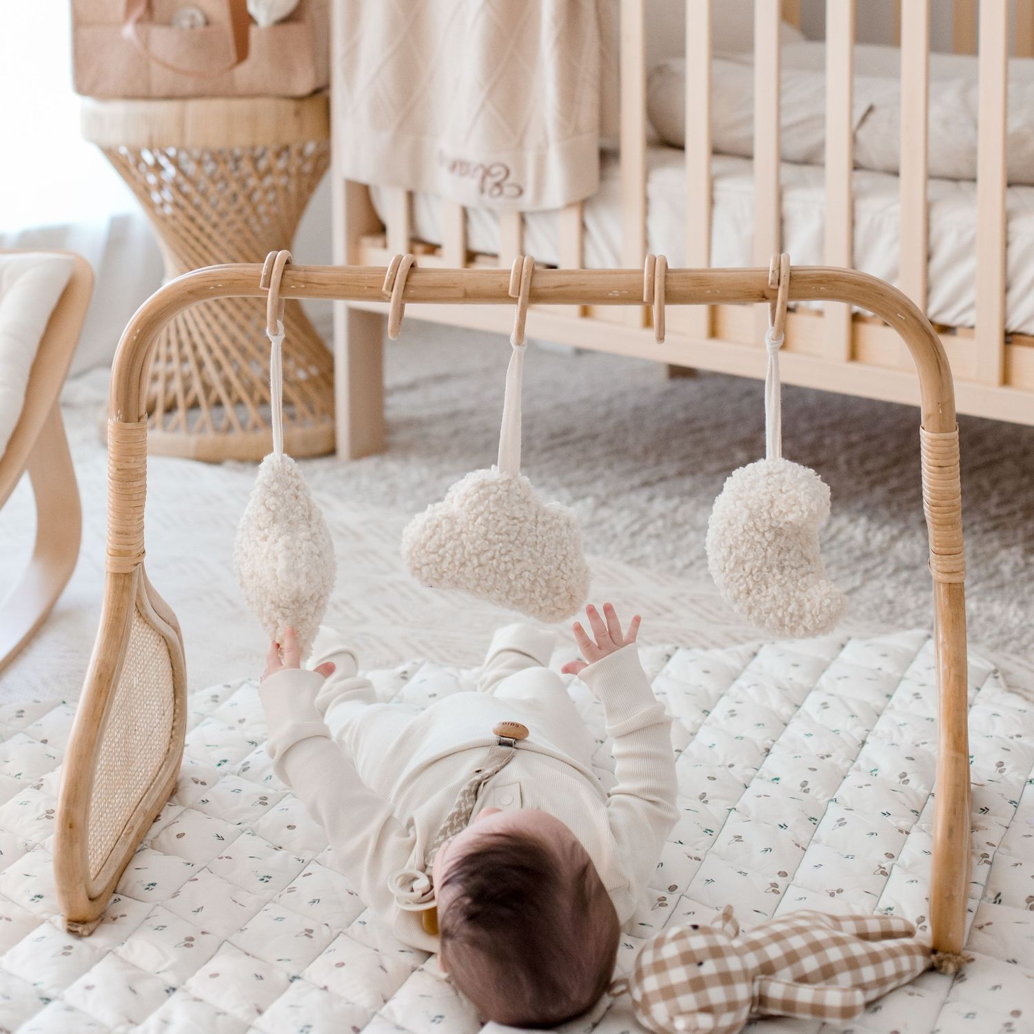 Baby lying under wooden play gym with hanging white pom poms in neutral nursery setting.