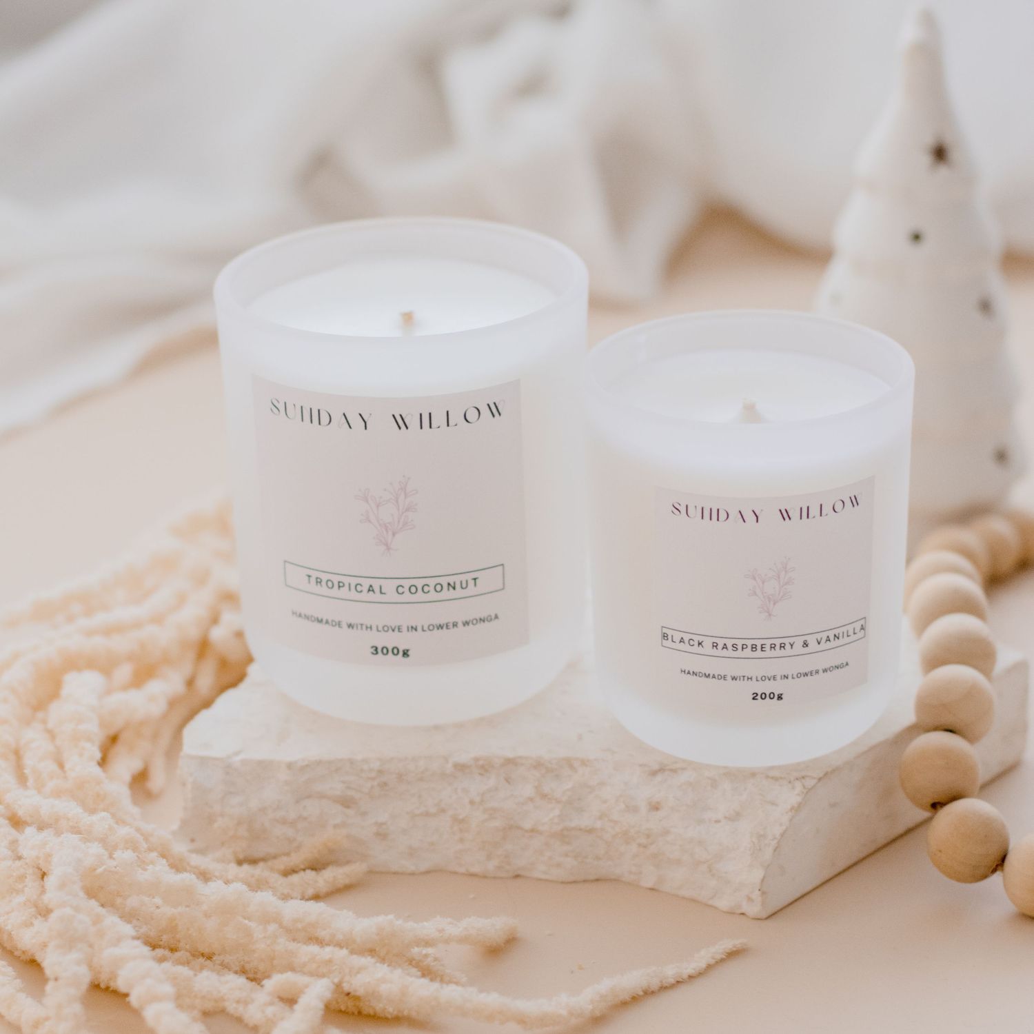 Two white Sunday Willow scented candles displayed on textured surface with wooden beads.