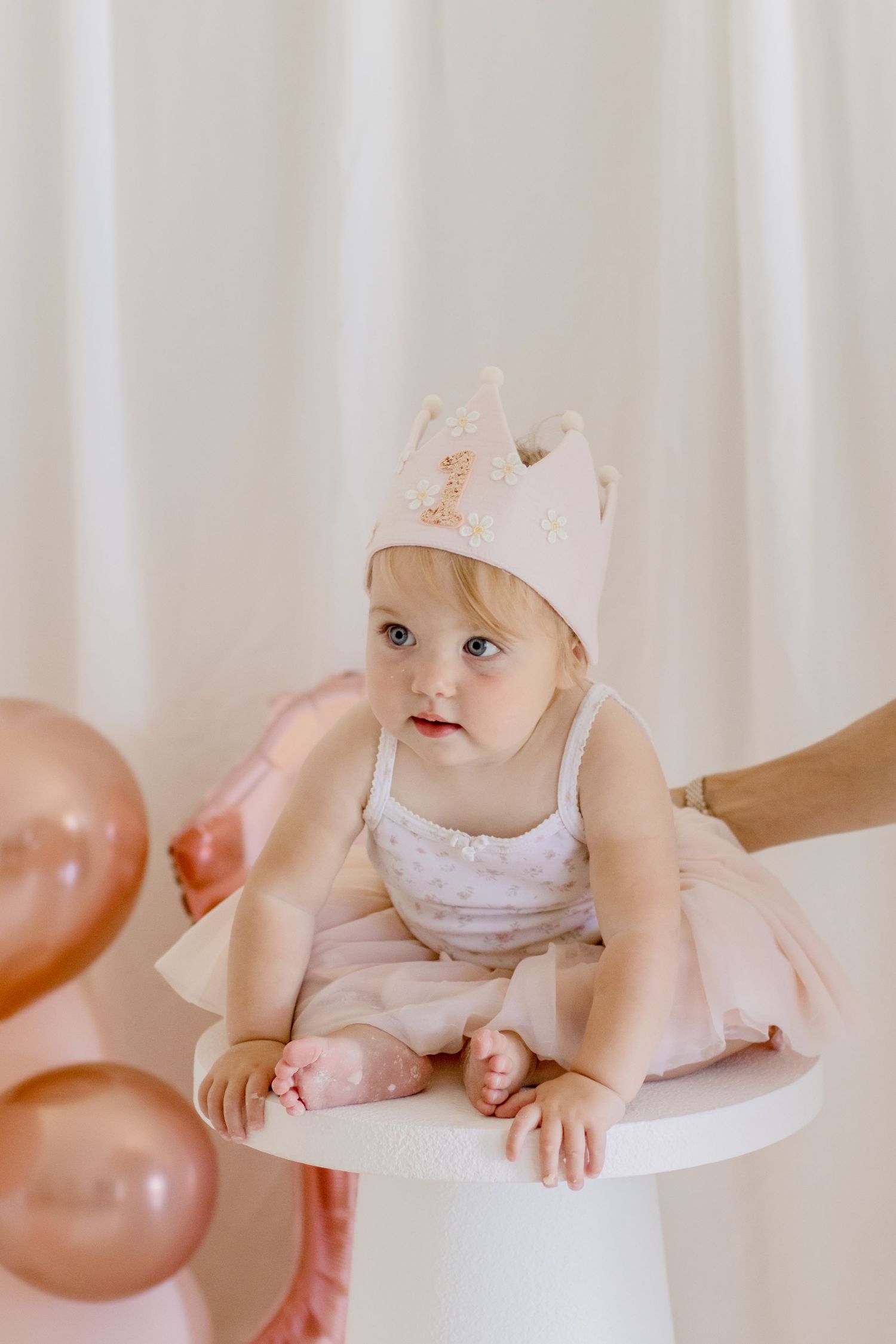 Baby in a pink ballet outfit and crown sits on white pedestal with rose gold balloons.
