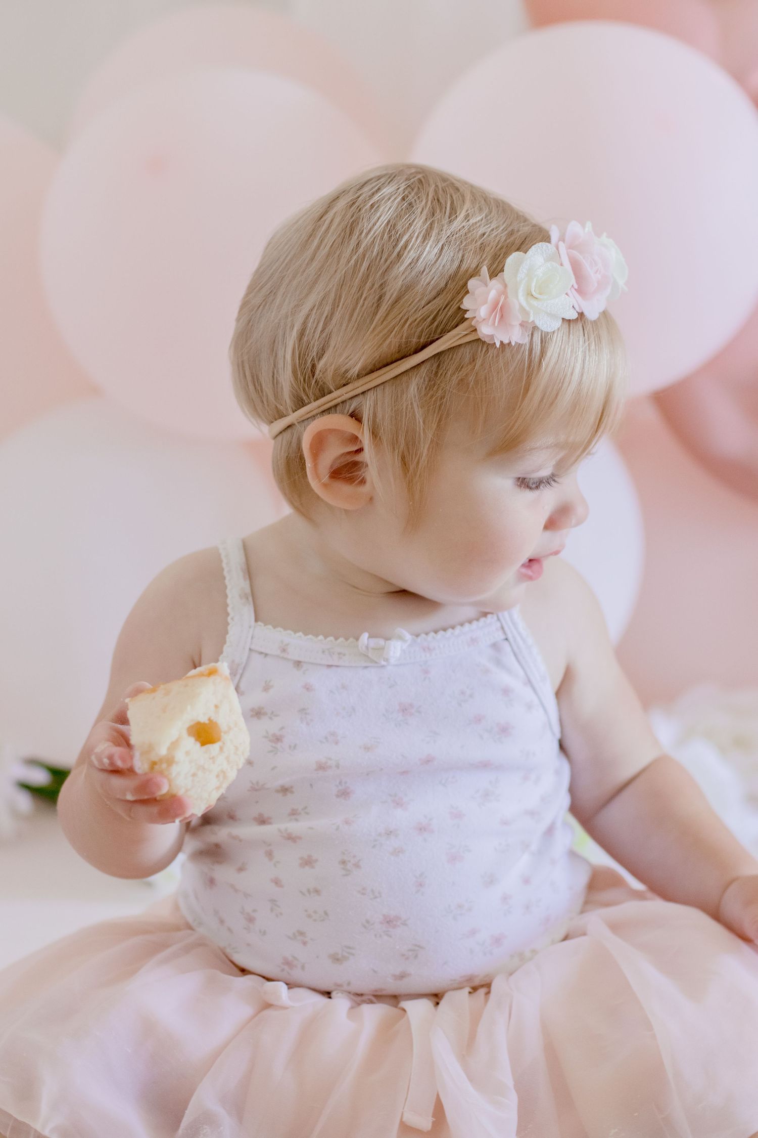 Baby in white dress with floral headband enjoying a snack against pink balloon backdrop.