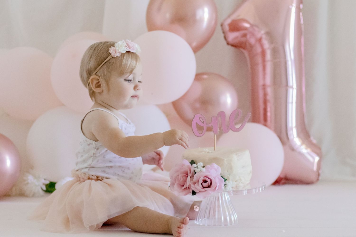 Baby in tulle dress playing with birthday decorations against pink balloon backdrop.