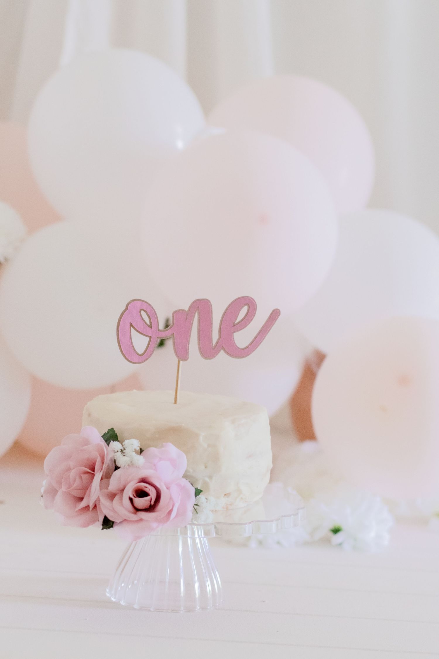 White birthday cake with pink 'one' topper and roses on glass stand against balloon backdrop.